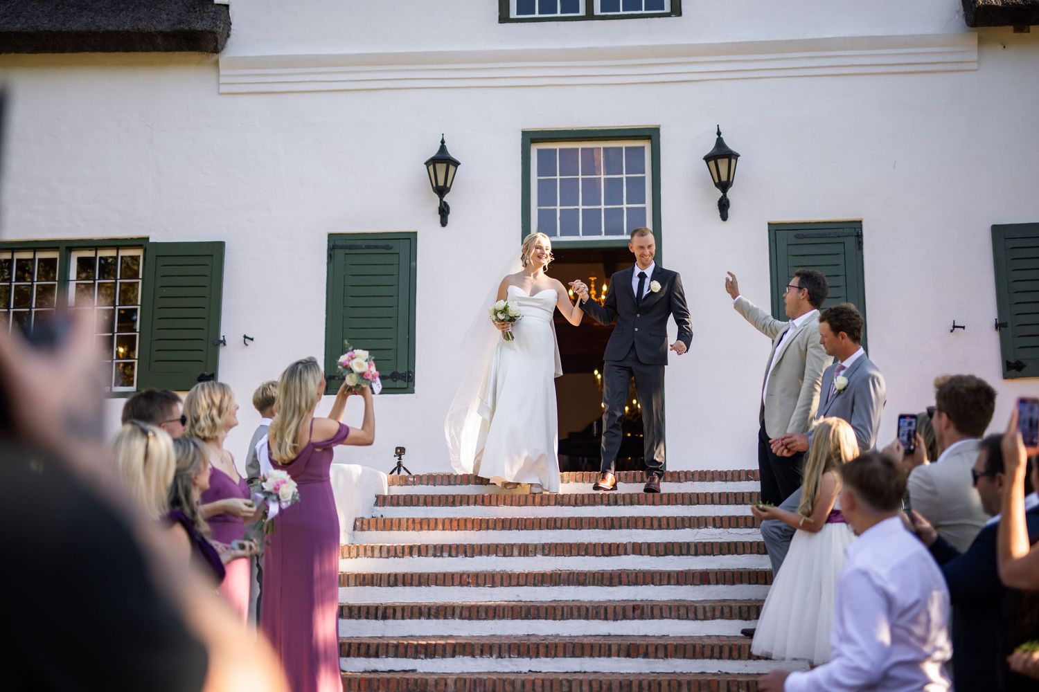 Wedding party gathers on white steps of historic building with green shutters.
