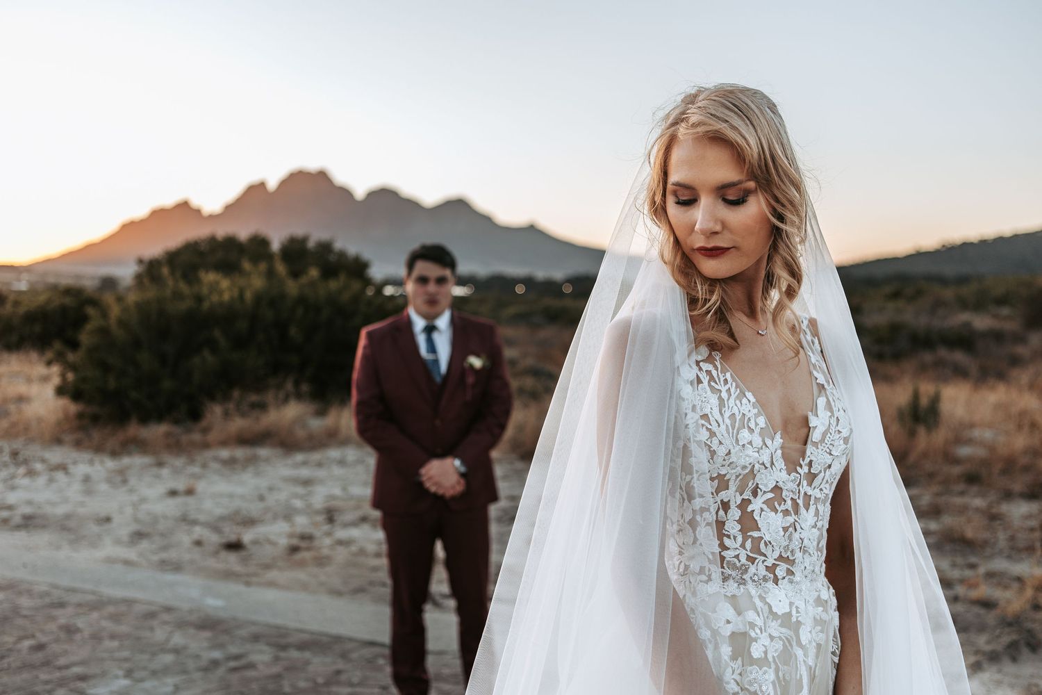 Elegant bride in lace gown with flowing veil against mountain backdrop at sunset.