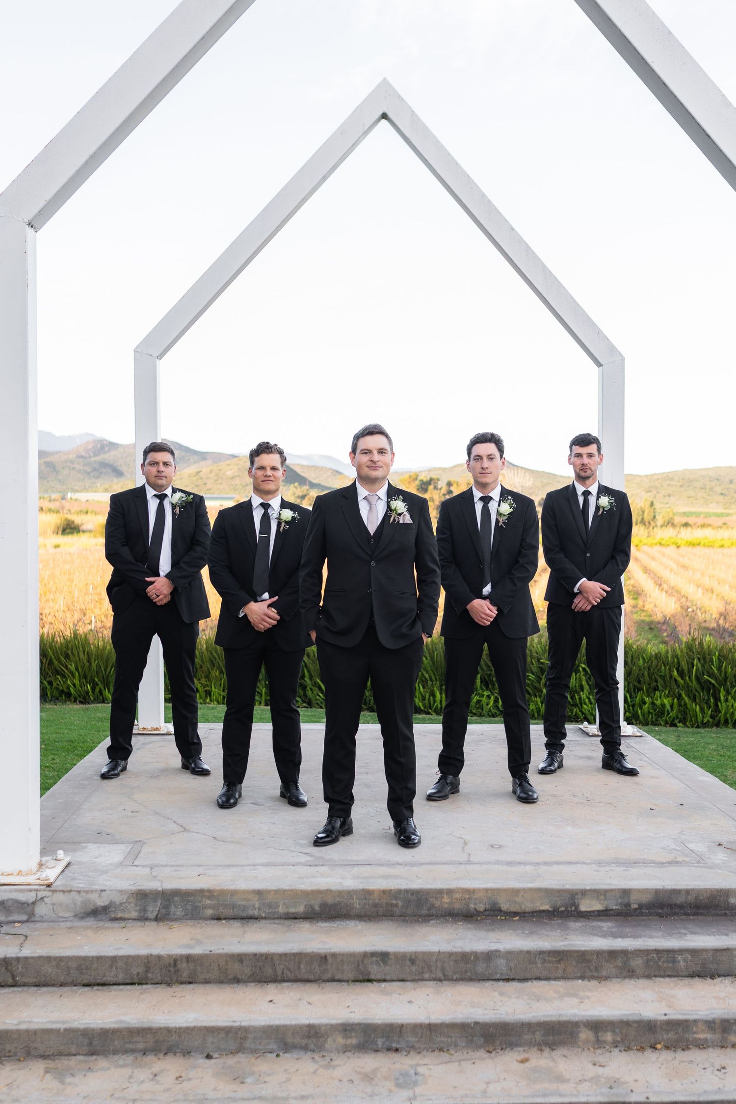 Groom and four groomsmen in black suits posing under white arch with mountain backdrop.
