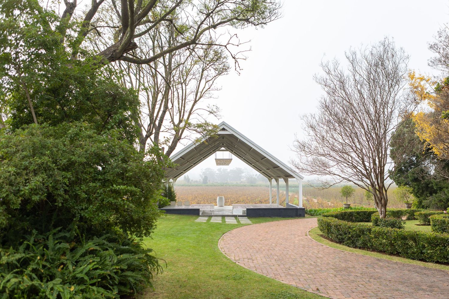 A serene garden gazebo surrounded by lush greenery, hedges, and bare trees on a misty day.