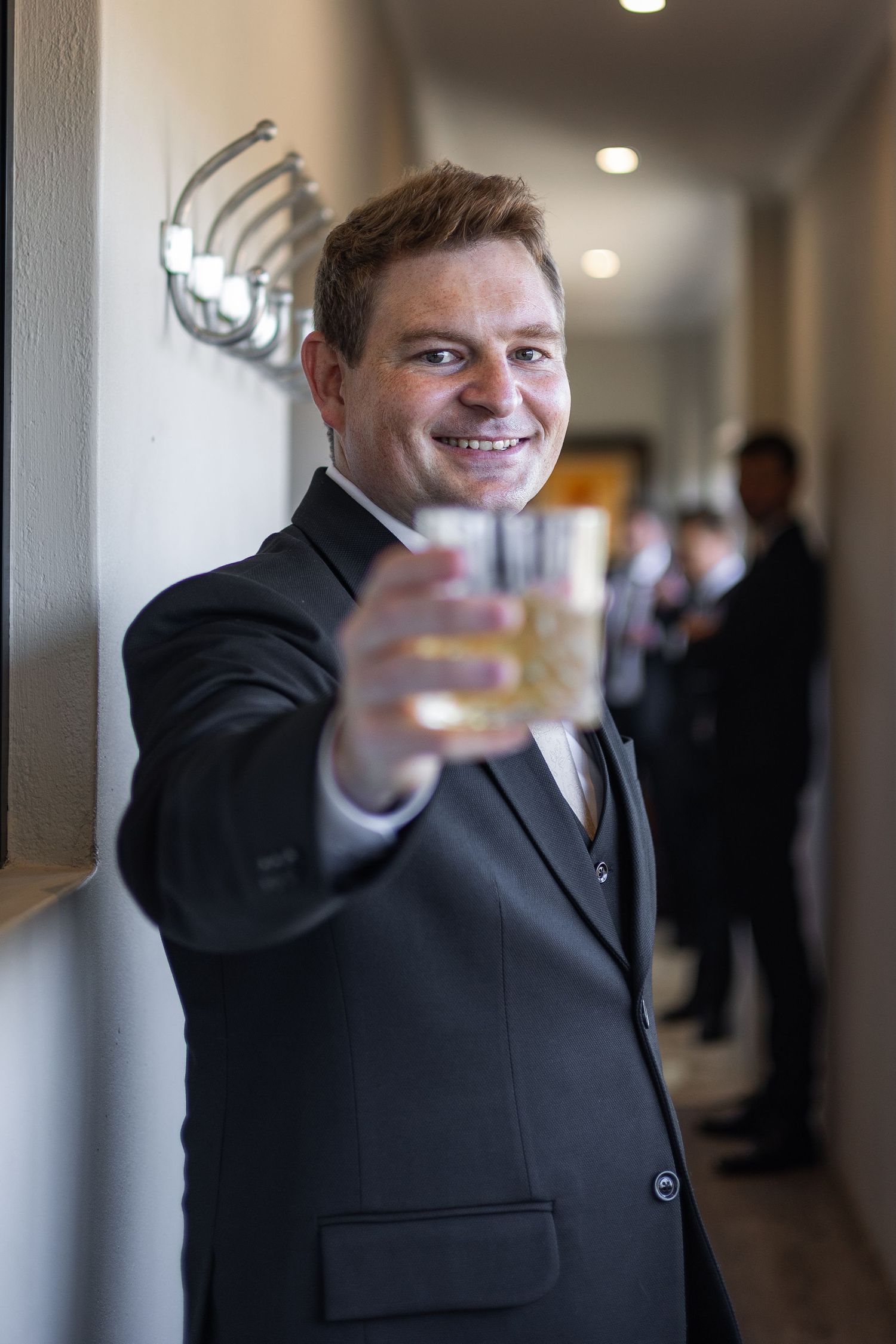Smiling groom in dark suit raising a whiskey glass toast before his wedding.