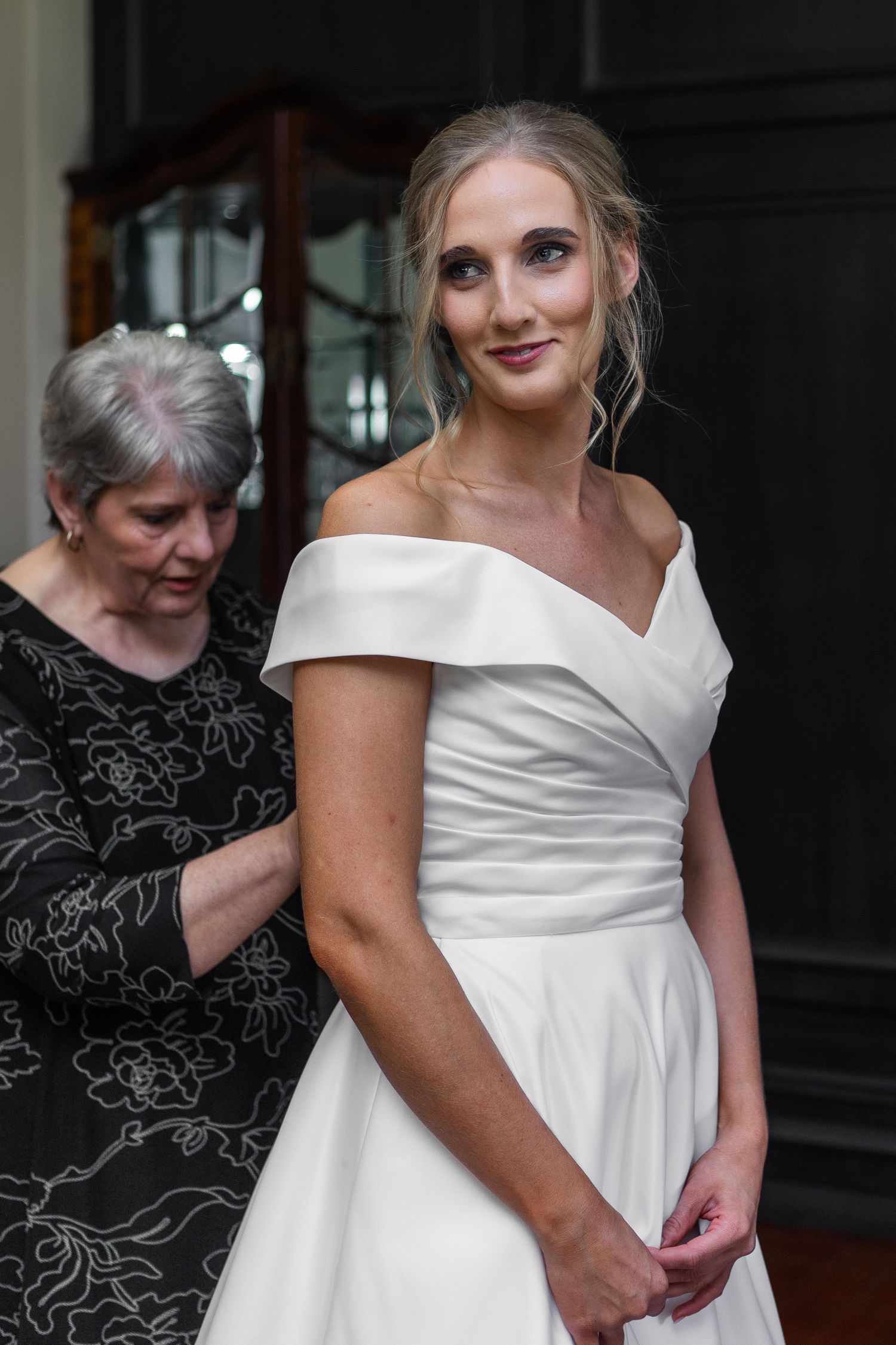 Older woman helping blonde bride fasten her off-shoulder white wedding gown from behind.
