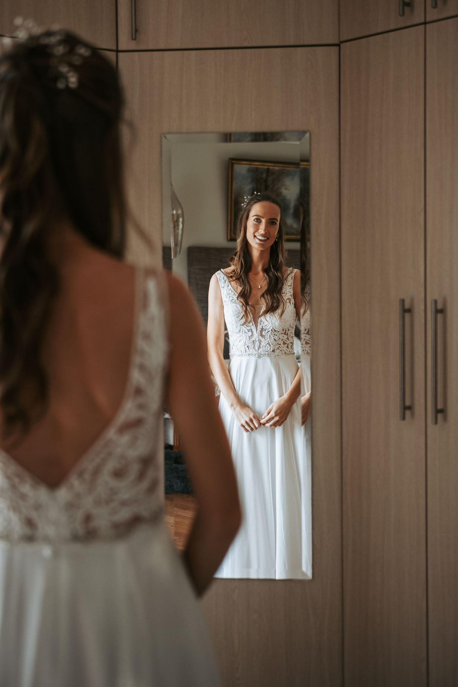 White lace wedding dress hangs in wardrobe mirror reflection during bridal preparation.