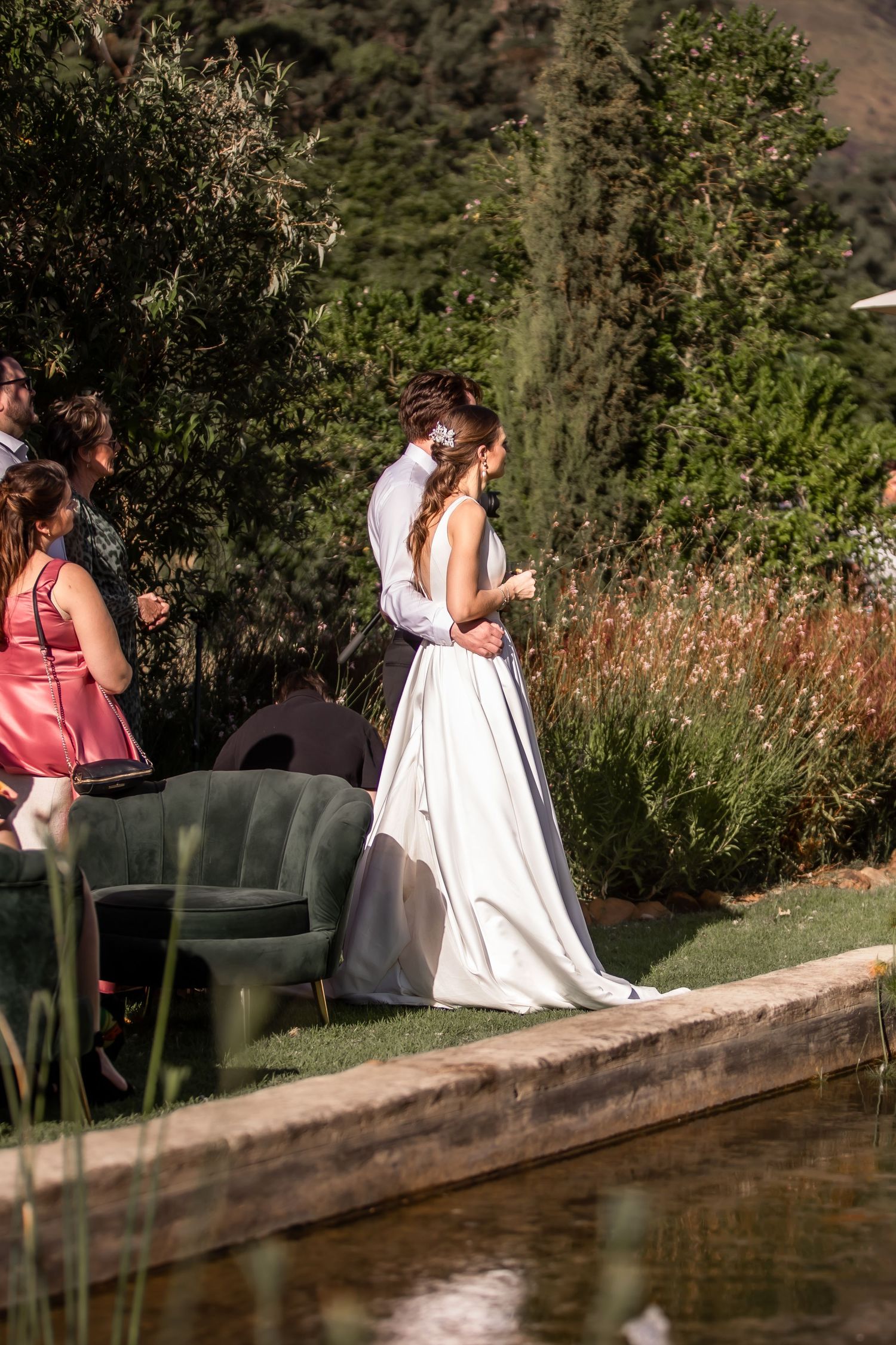 A bride in a white dress stands near a pond surrounded by lush greenery during an outdoor wedding ceremony.