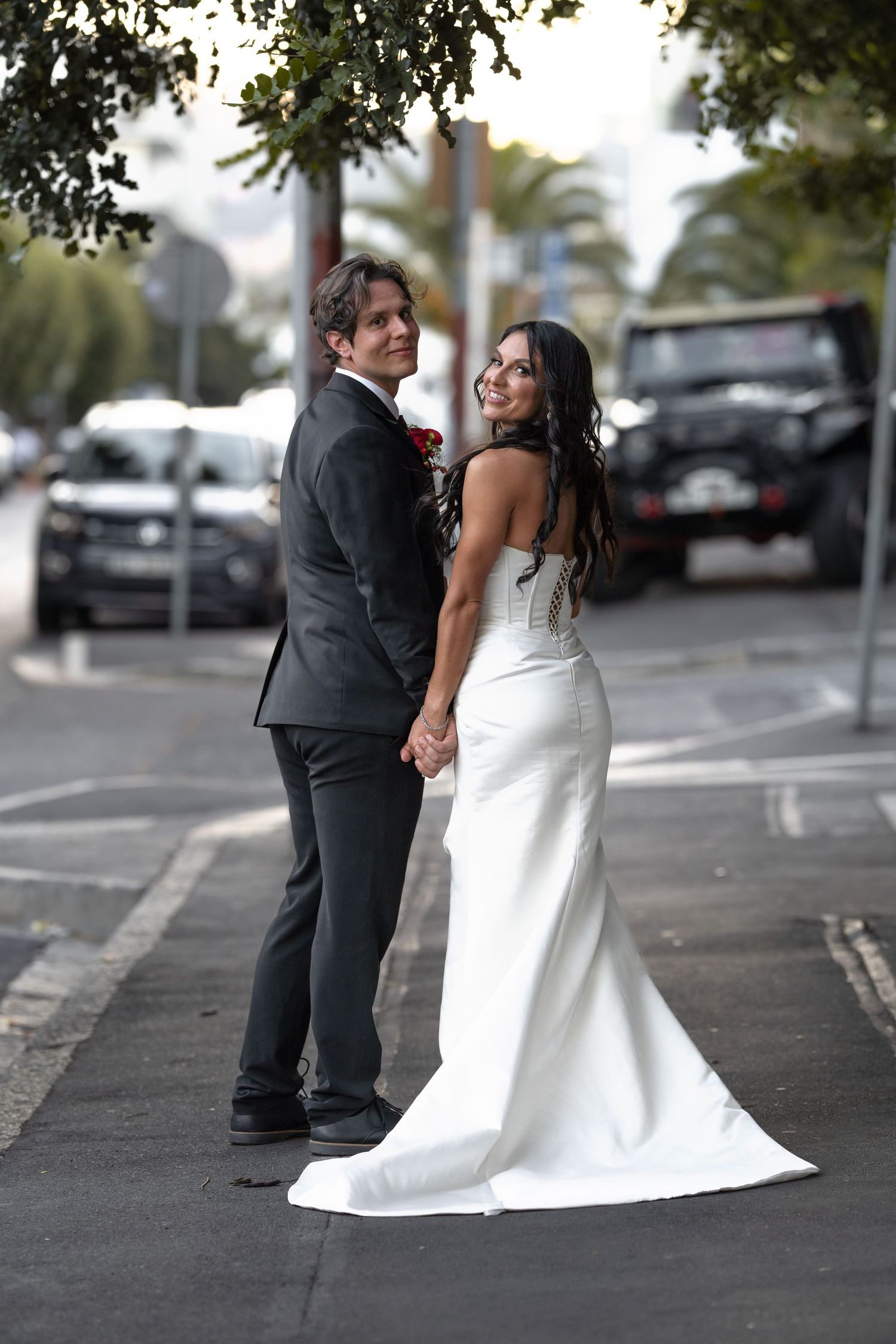 Bride in white gown and groom in gray suit hold hands on a sunlit street, both smiling over their shoulders.