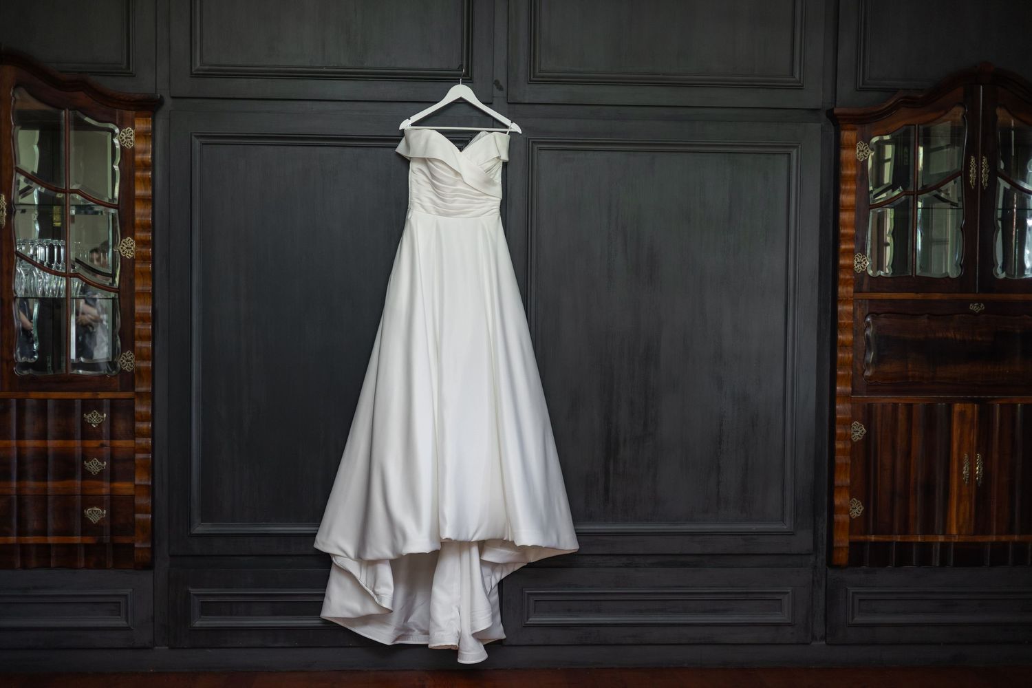 Elegant white off-shoulder wedding gown hanging on a hanger against a dark gray wood-paneled wall.