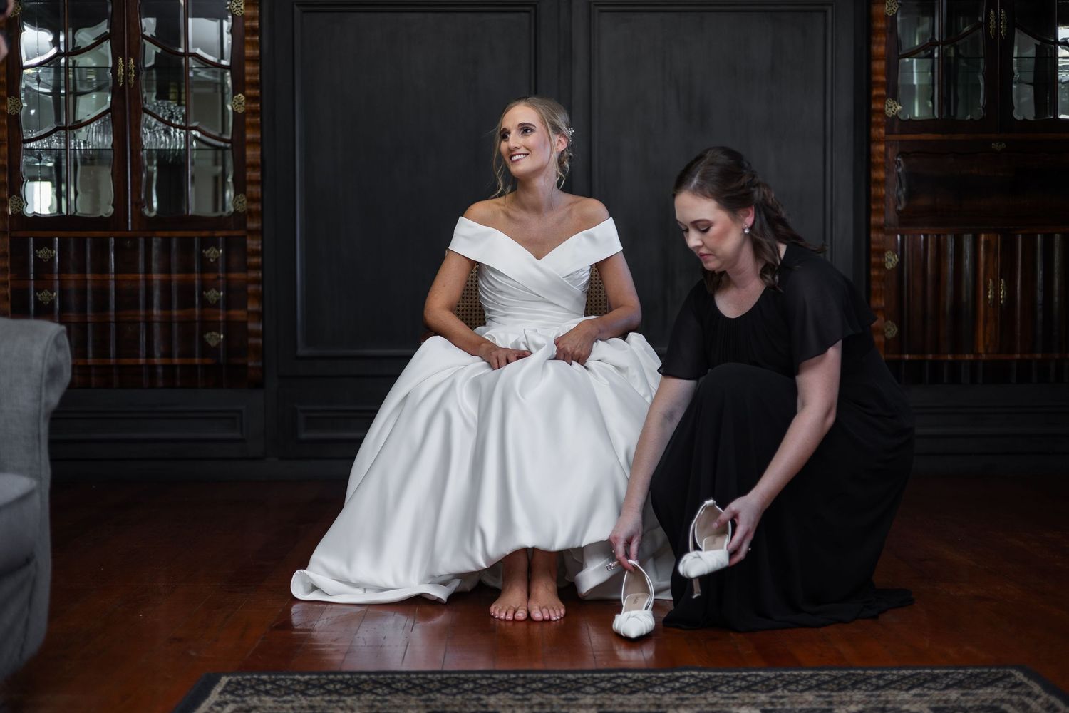 Bride in white off-shoulder gown being helped with shoes by bridesmaid in black dress.