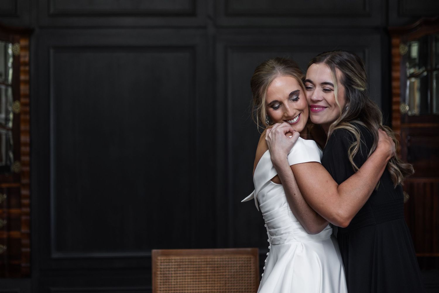 Bride in white dress shares a warm embrace with a bridesmaid in a black dress.