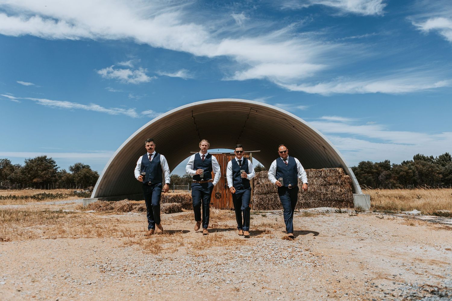 Wedding party stands in line against curved metal building on sunny day with blue skies.