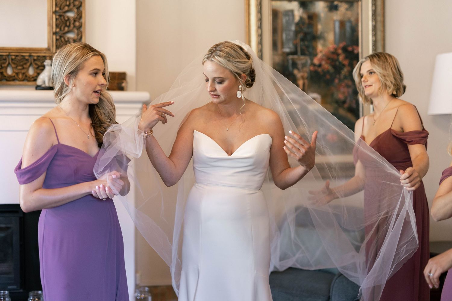 Wedding party in purple bridesmaid dresses helps bride get ready in elegant dressing room.