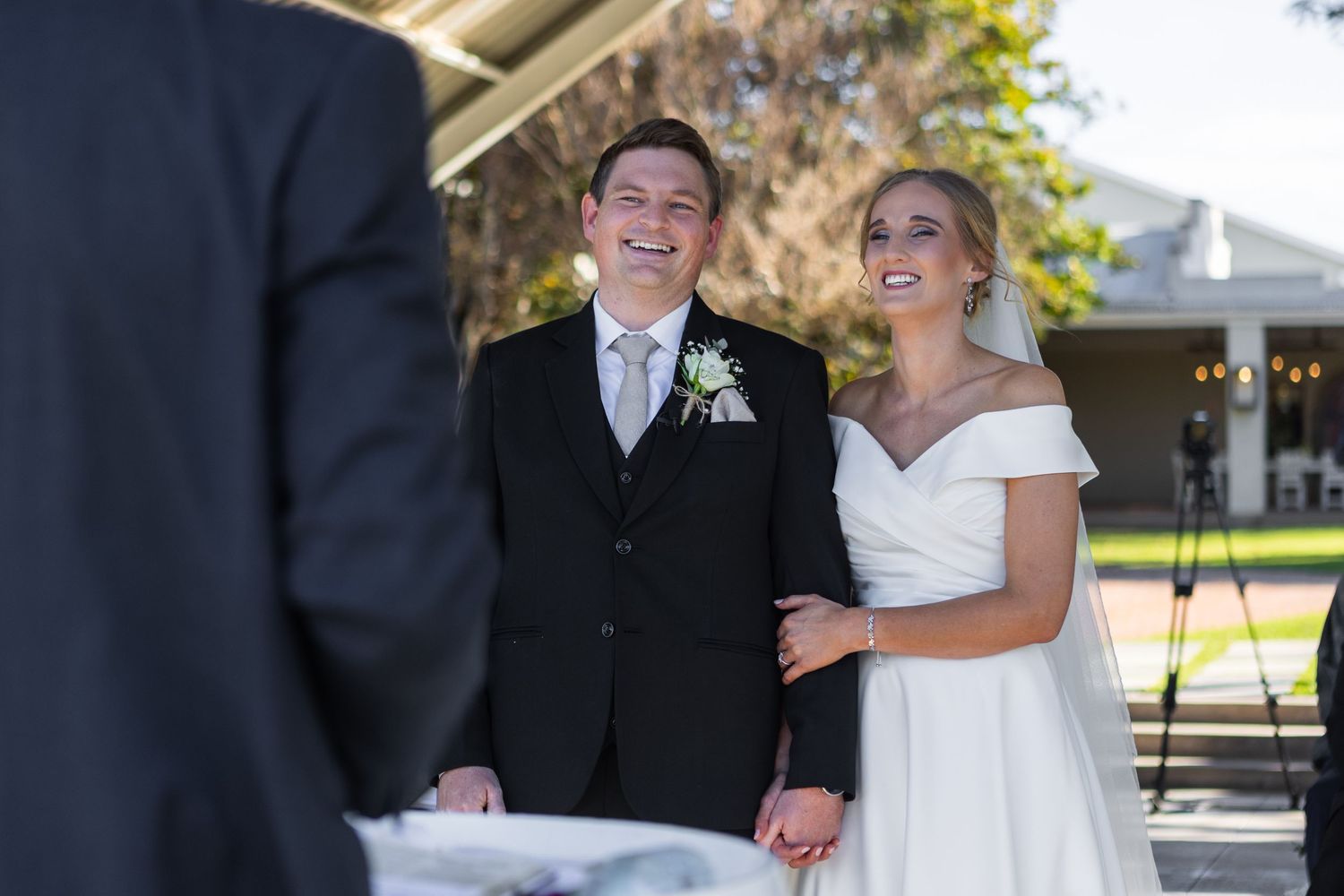 Smiling bride and groom holding hands during outdoor wedding ceremony with officiant.