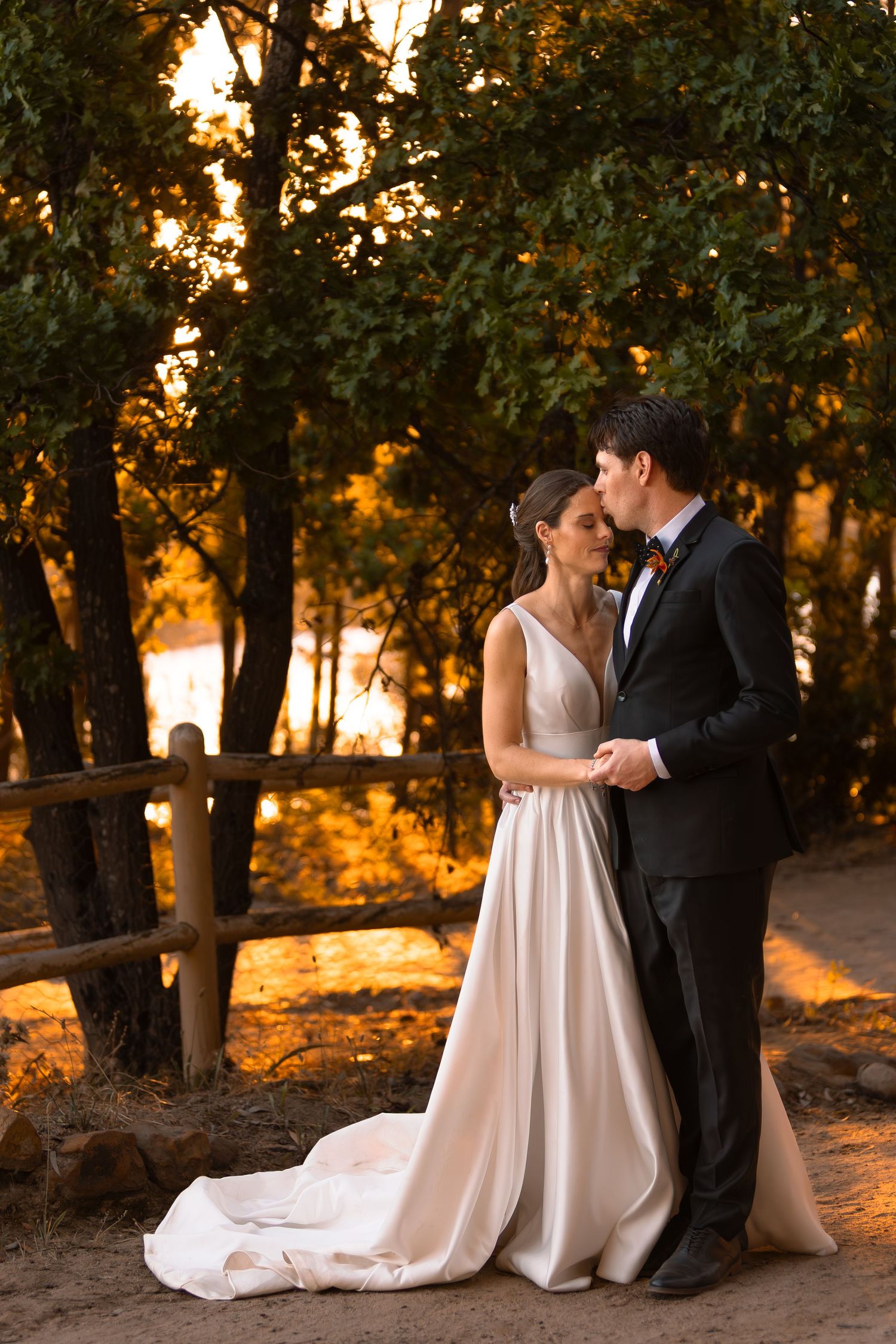 Romantic wedding couple poses in a wooded area during golden hour sunset with flowing white dress and black suit.