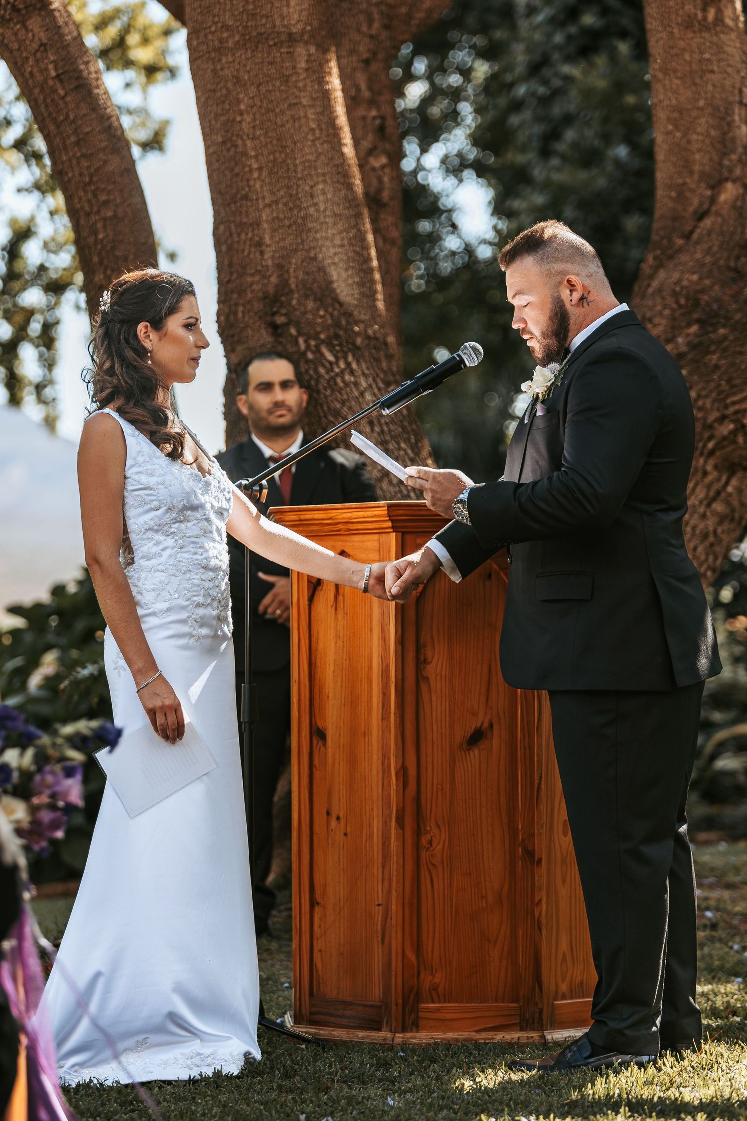 Wedding ceremony taking place under large trees with wooden podium and guests in attendance.