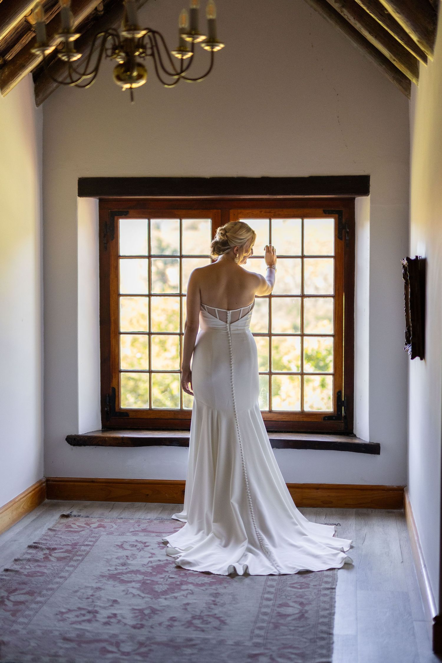 Bride in elegant white wedding gown with train stands before sunlit window in rustic chapel interior.