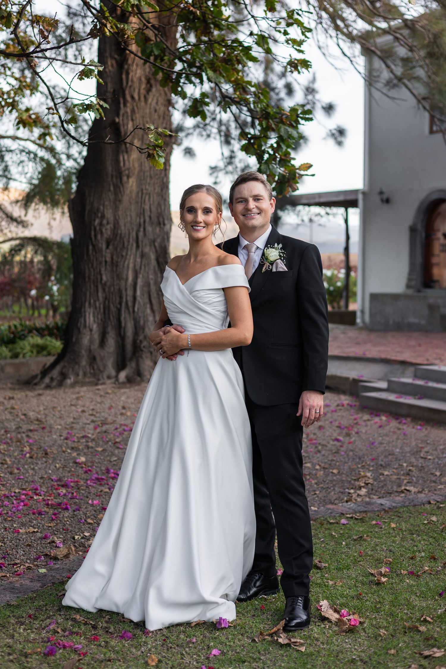 Bride and groom embracing under large tree with purple petals on ground.