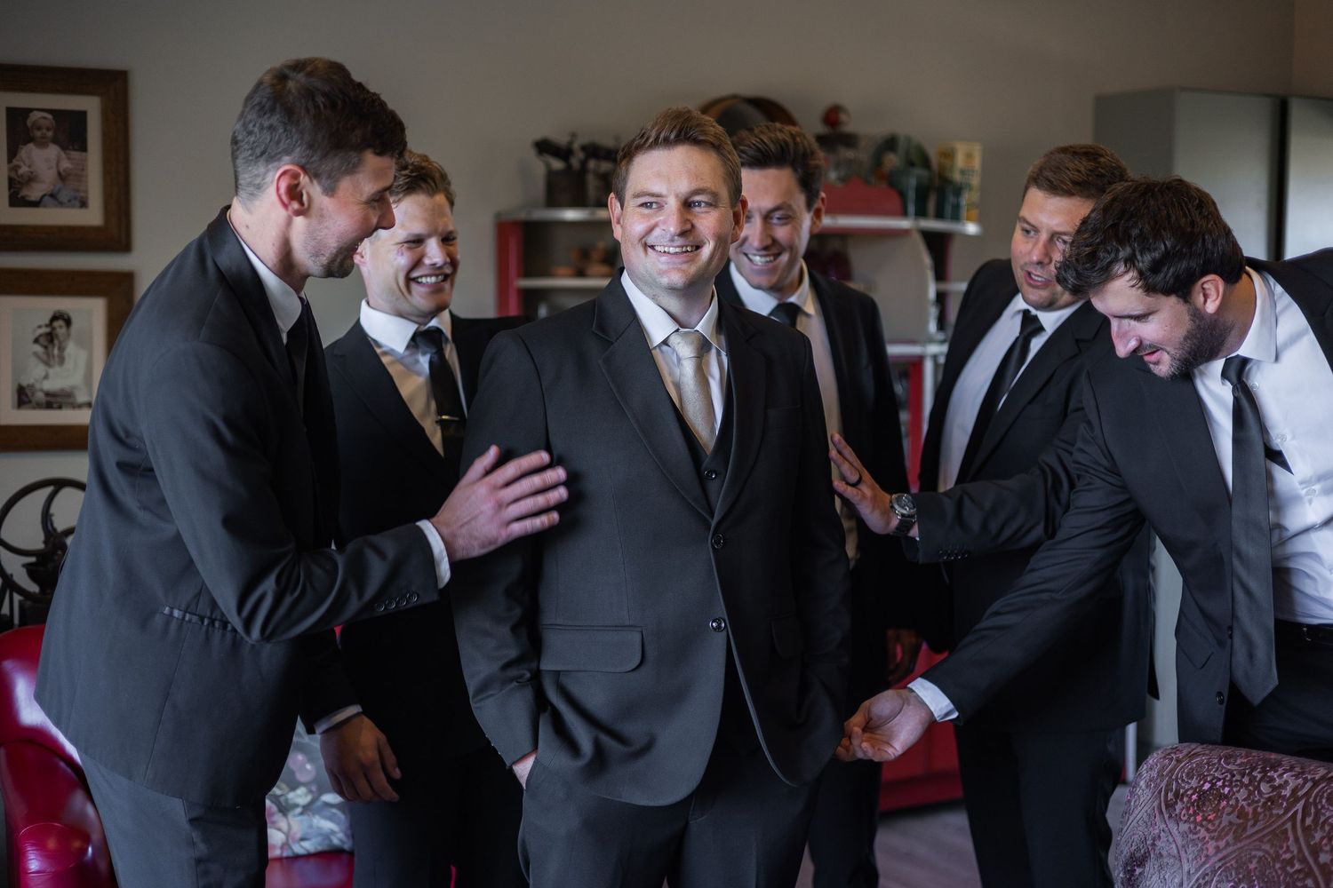 Groom surrounded by smiling groomsmen in dark suits during wedding preparations indoors.