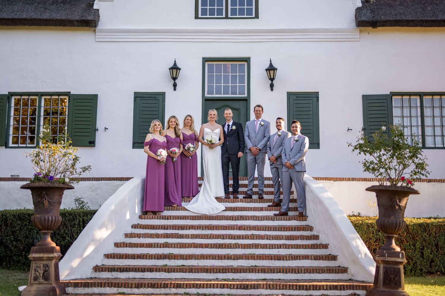 People gathered on white steps of a colonial style building with green shutters and lanterns on the exterior walls.