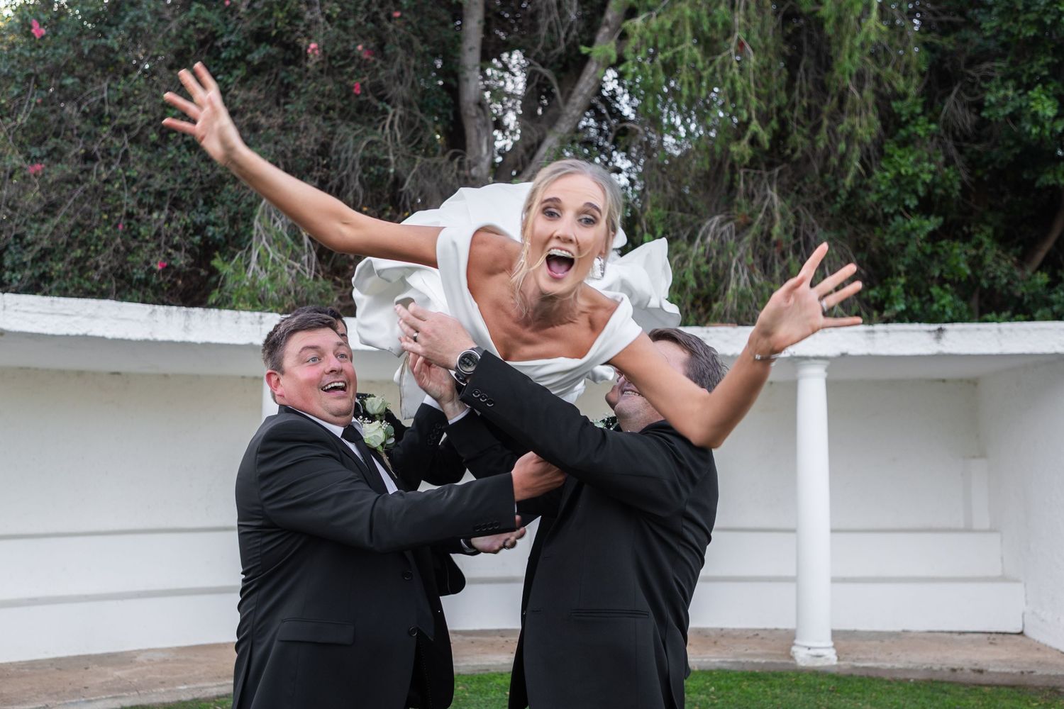 Joyful bride tossed in air by groomsmen in black suits during outdoor wedding celebration.