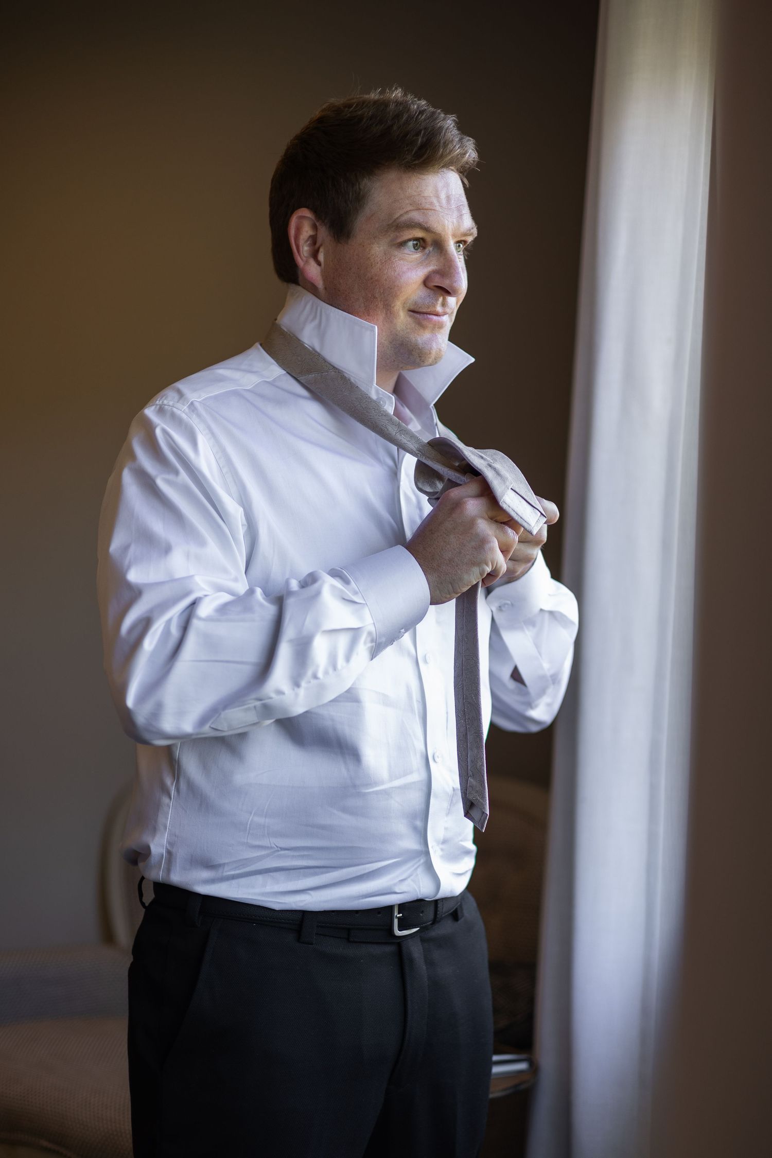 Elegant groom in white shirt adjusting gray tie while gazing out a sunlit window.
