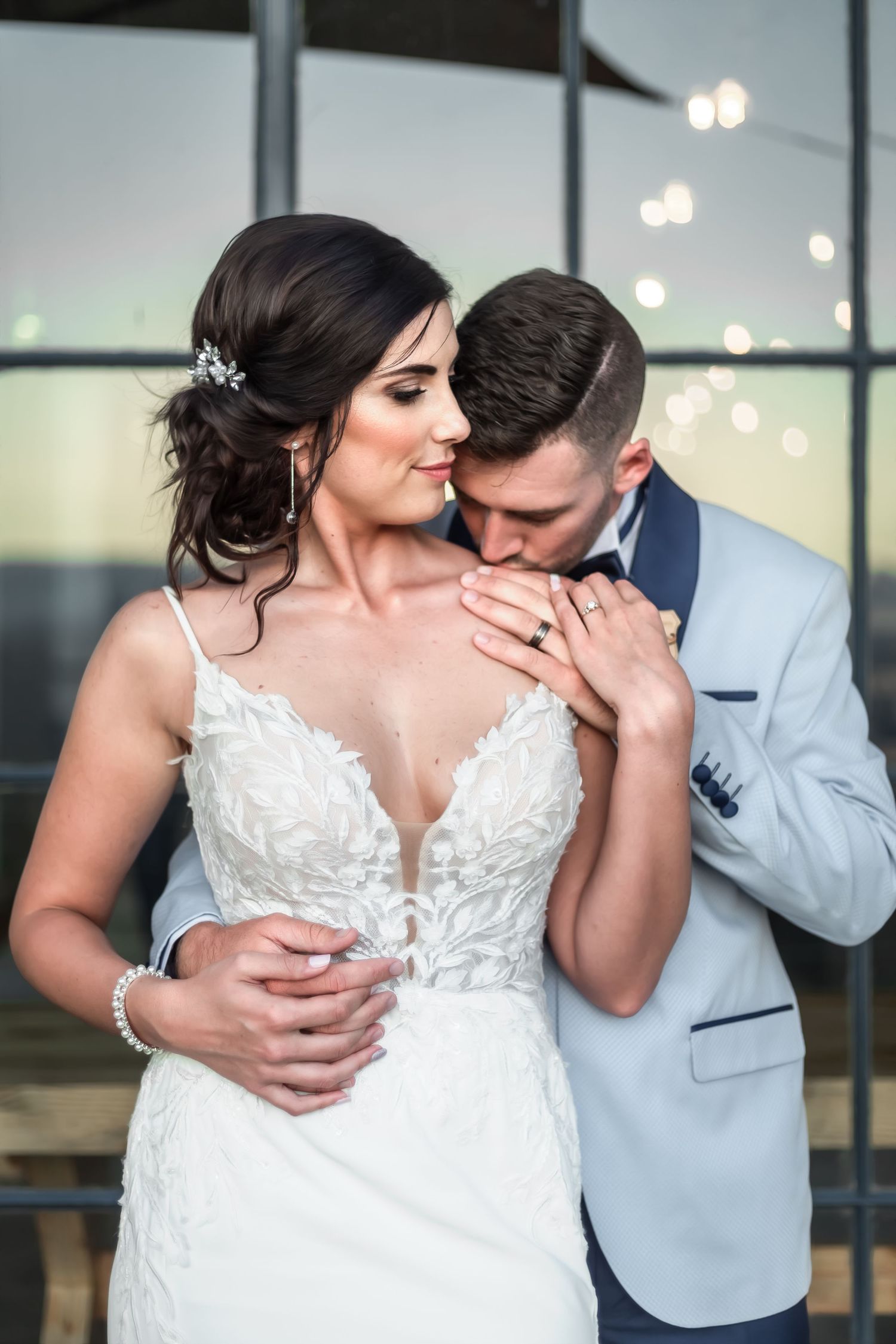 Romantic wedding couple shares intimate moment in elegant attire with soft lighting and modern interior backdrop.