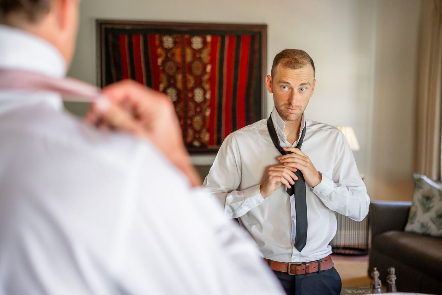 A groom adjusts his tie while getting ready in a room with decorative wall art.