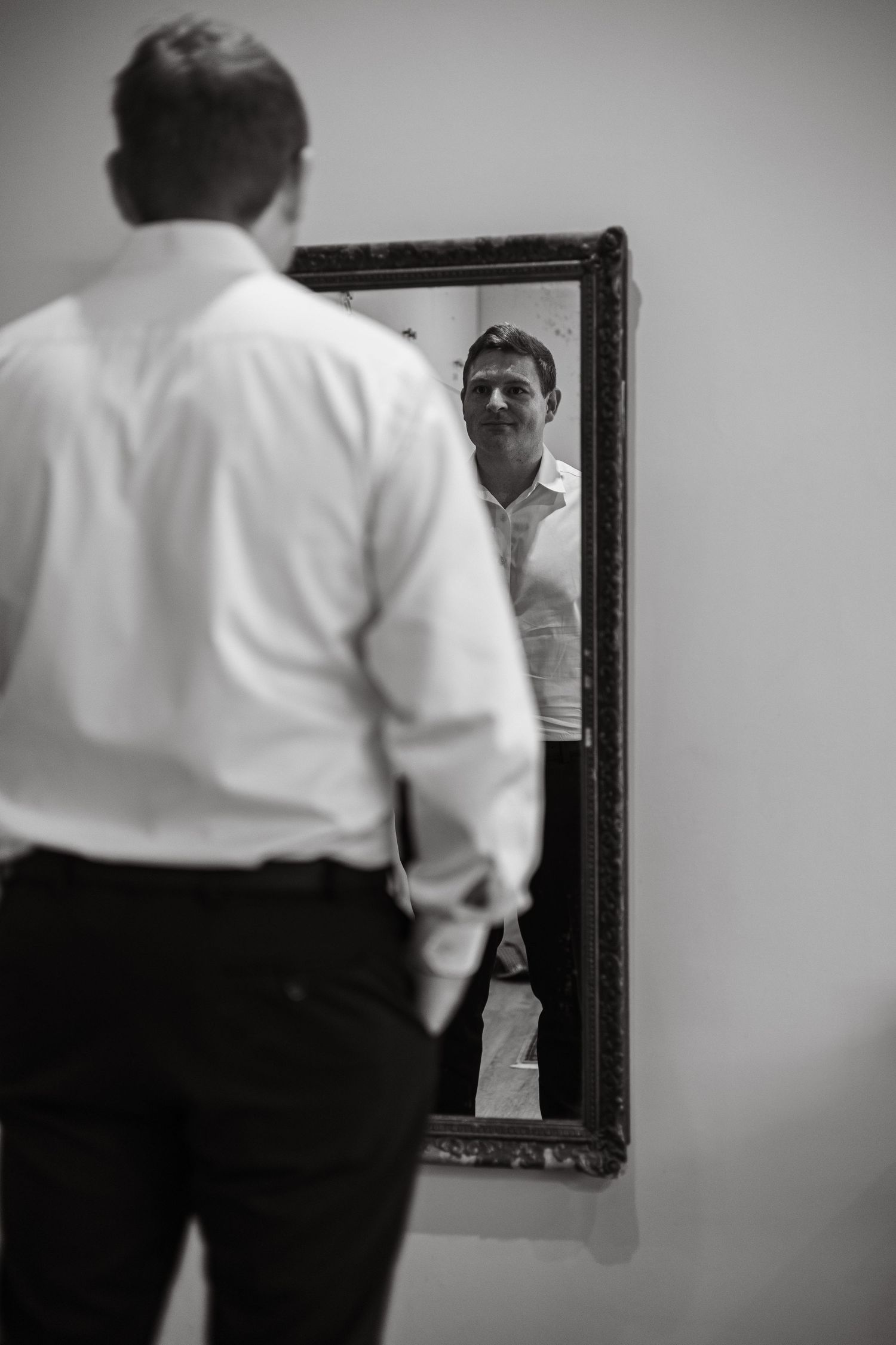 Groom in white shirt checks his appearance in an ornate mirror while preparing for his wedding day.