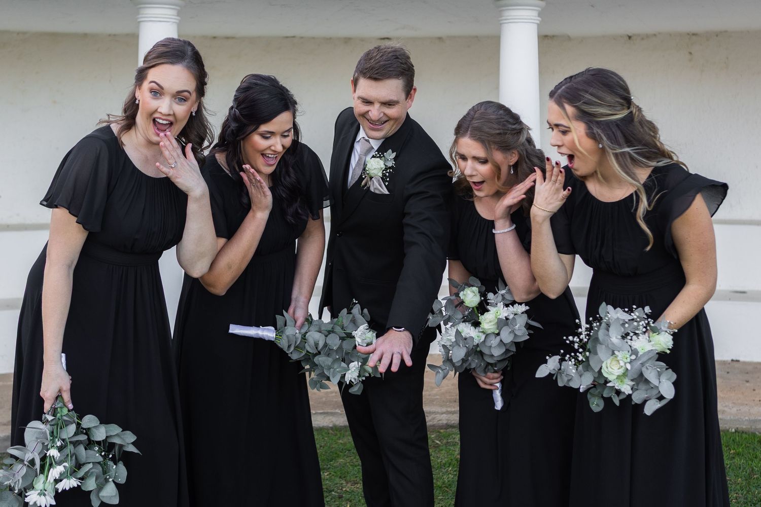 Bridal party in black dresses and suit laughing together outdoors holding white floral bouquets.