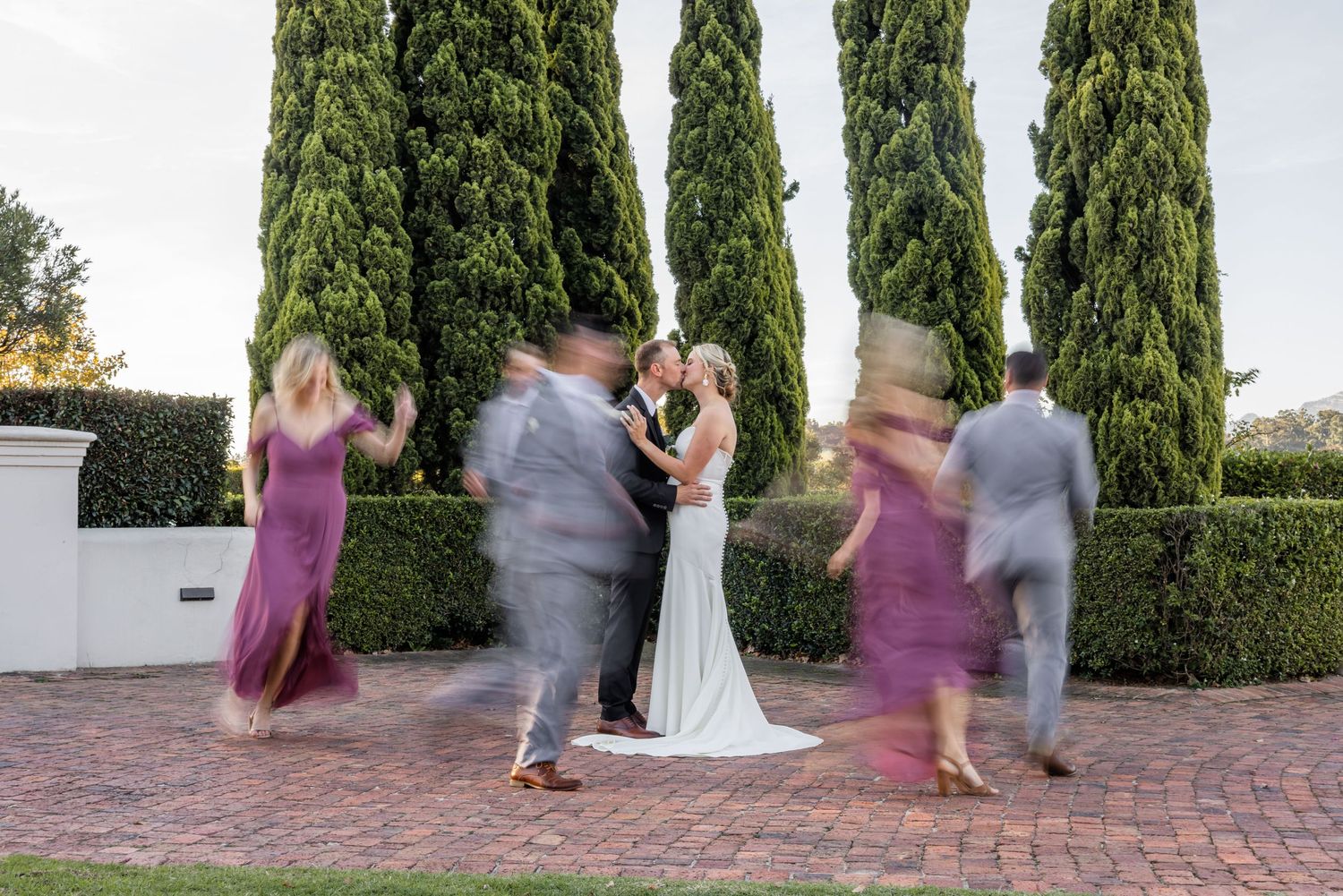 Bride and groom kiss surrounded by blurred dancing wedding party guests near tall cypress trees on brick patio.