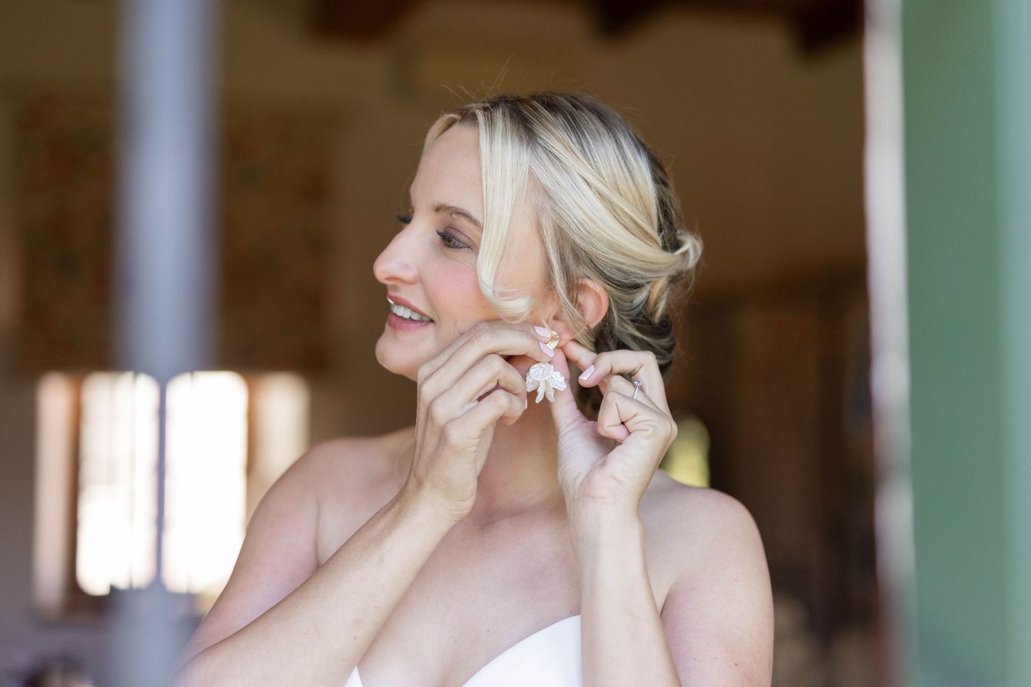 A person wearing a white dress adjusts earrings while looking in a mirror with a soft smile.