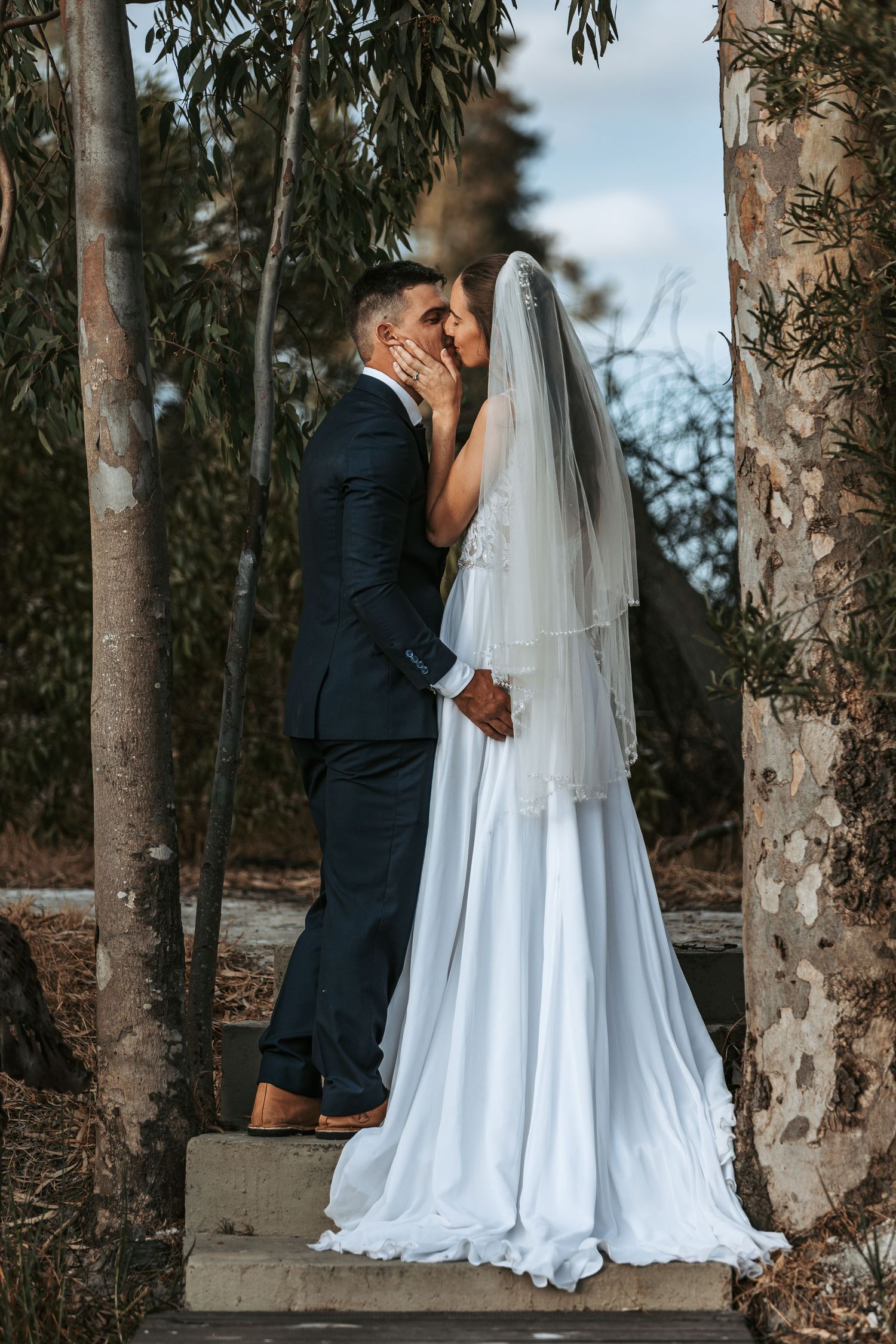 Couple in wedding attire embraces on stone steps surrounded by tall trees.