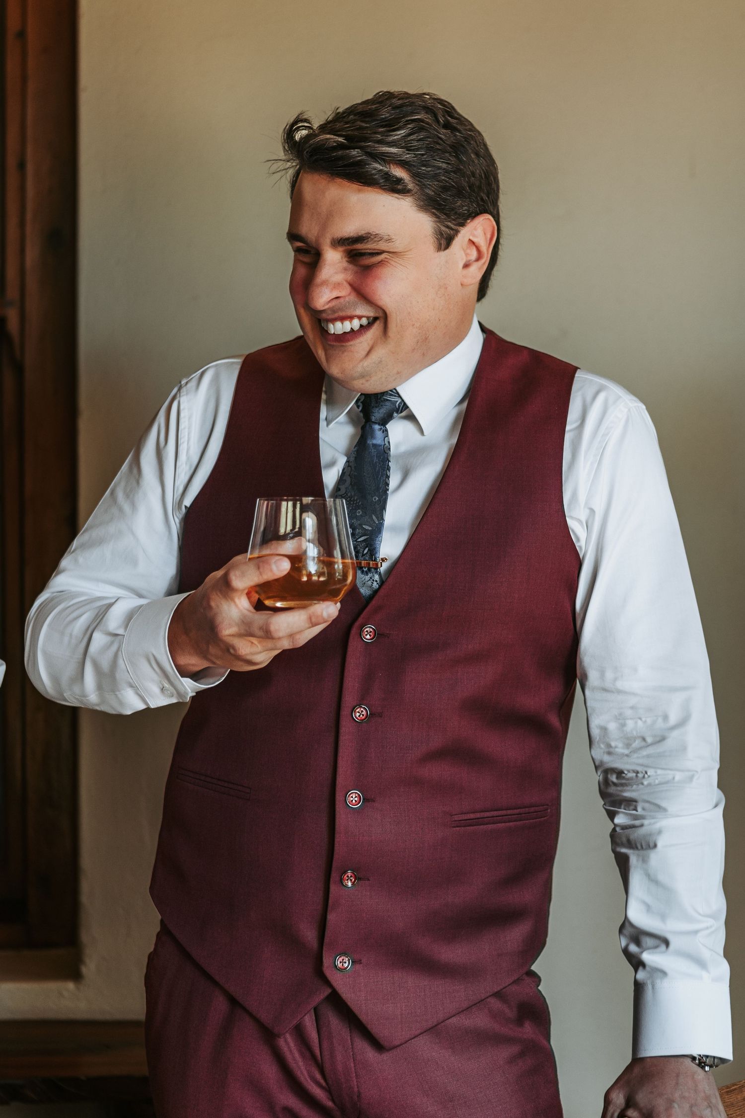Wedding party member in burgundy vest smiles while holding glass of spirits during preparations.