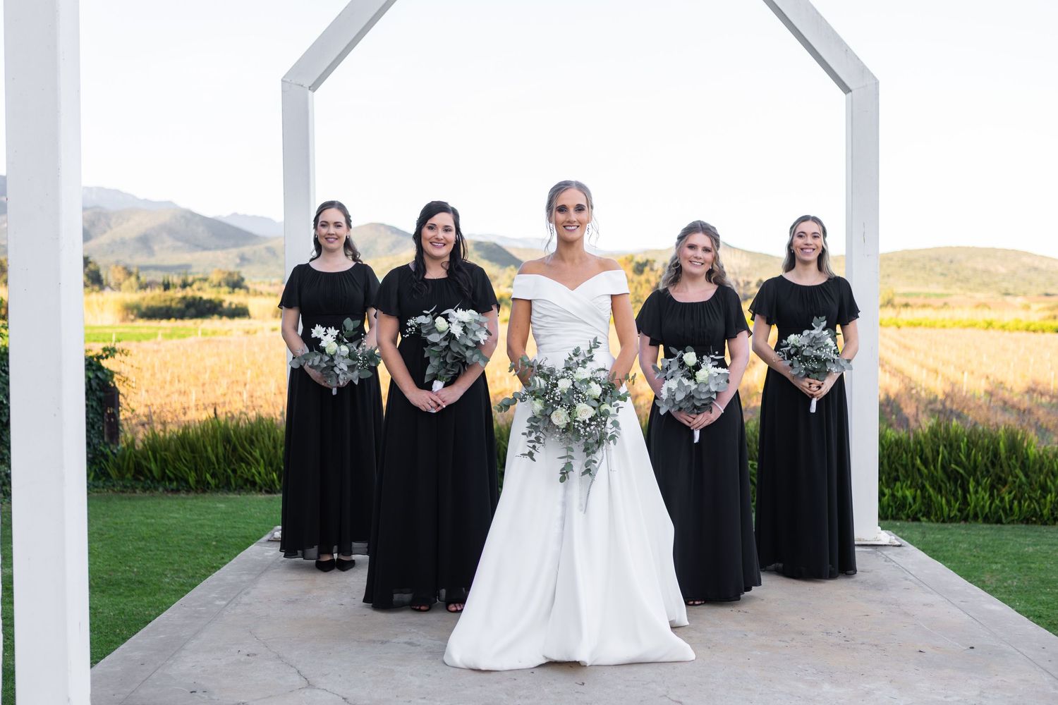 Bride in off-shoulder white gown poses with four bridesmaids in black dresses holding bouquets at a vineyard.