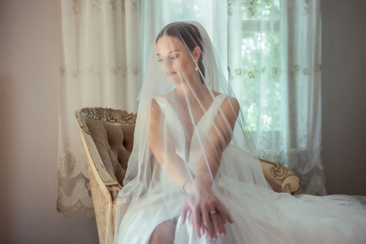 Ethereal bridal boudoir photo with flowing white veil and elegant vintage chair in natural window light.