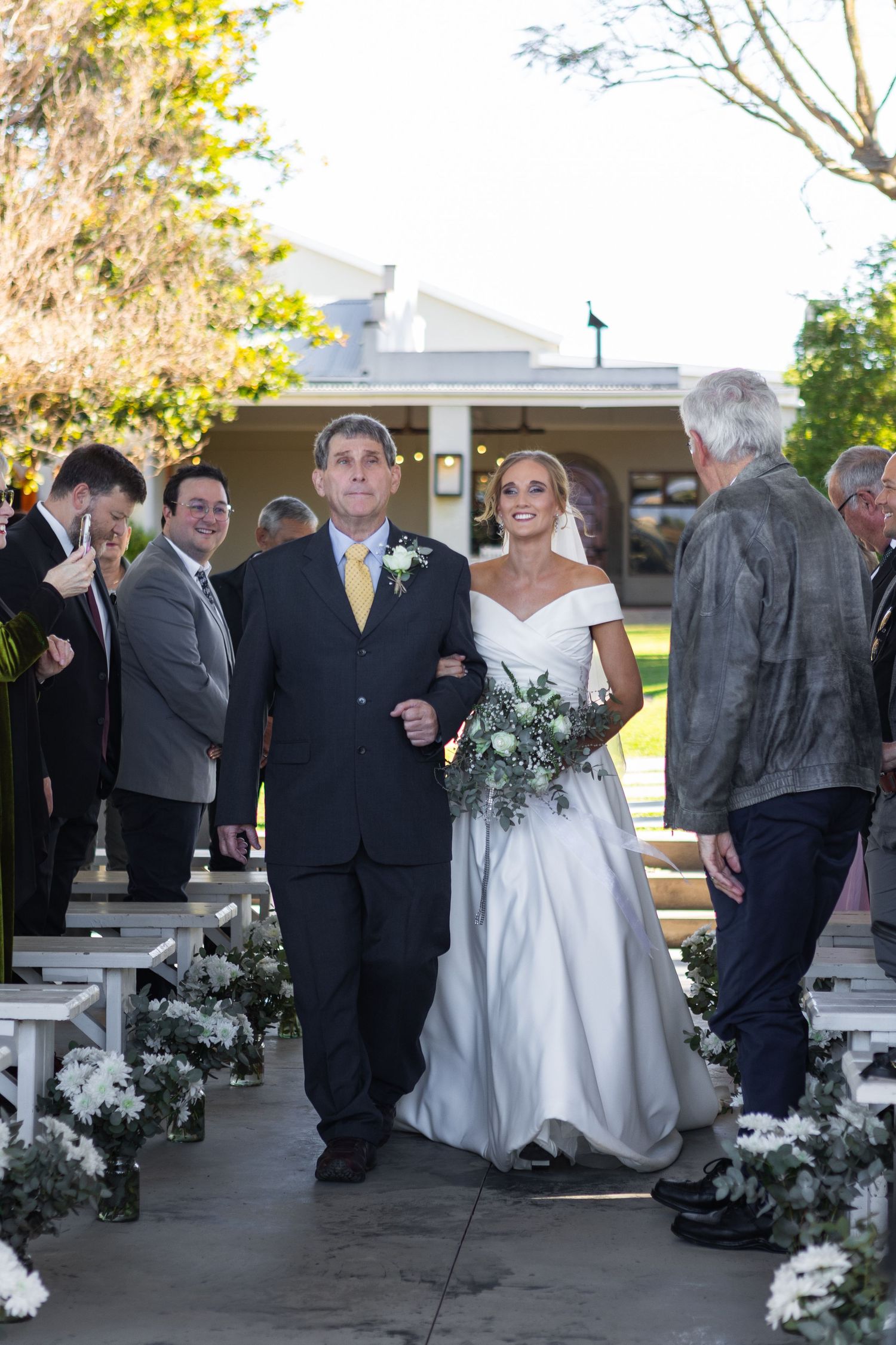 Bride in white off-shoulder gown walking down the aisle with her father outdoors.
