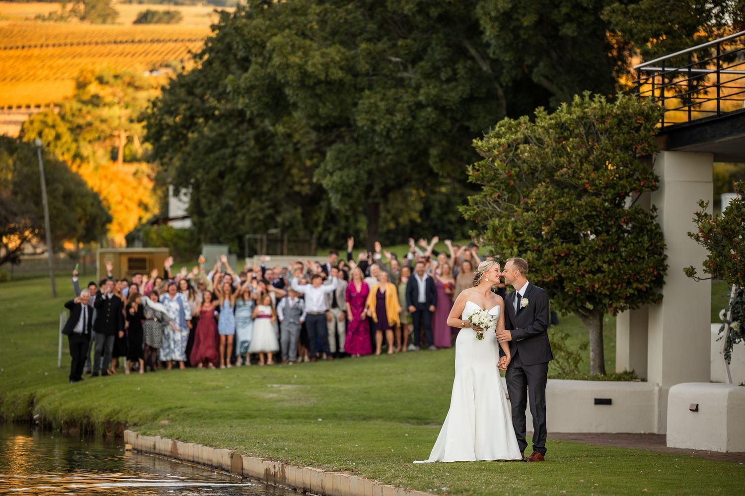 Wedding guests gather outdoors on a lush green lawn during golden hour at an elegant garden ceremony.