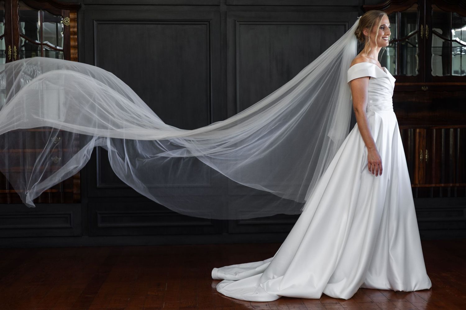 Bride in off-shoulder white gown with flowing cathedral veil in elegant dark wood interior.