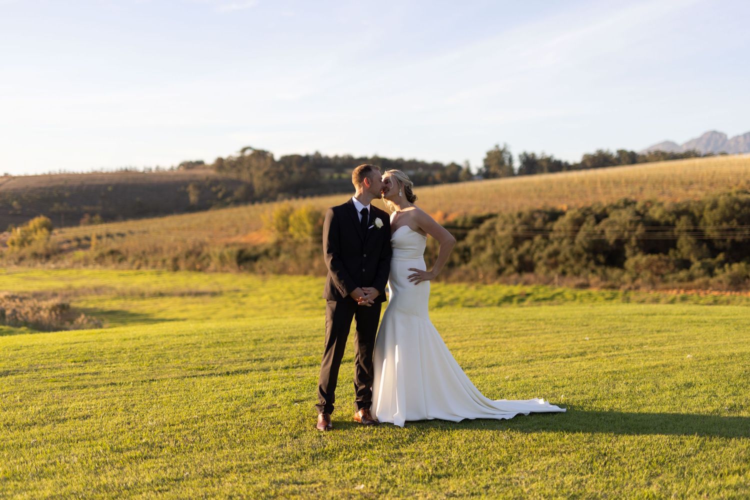 Newlywed couple walking together across a scenic hilltop meadow at sunset.
