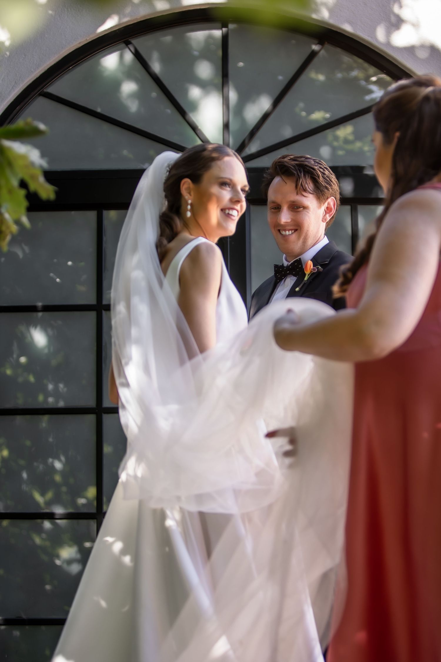 A wedding couple shares a joyful moment under an arched window with flowing veil and elegant attire.
