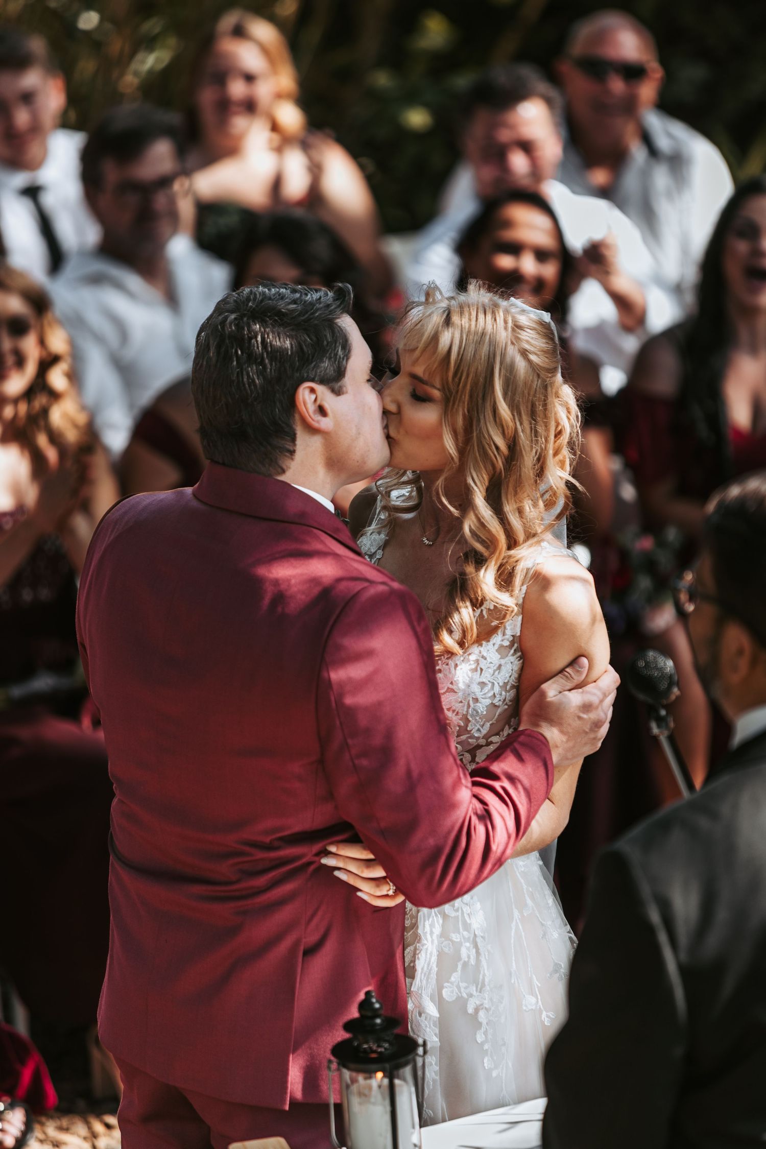 A joyful first kiss at an outdoor wedding ceremony surrounded by applauding guests.