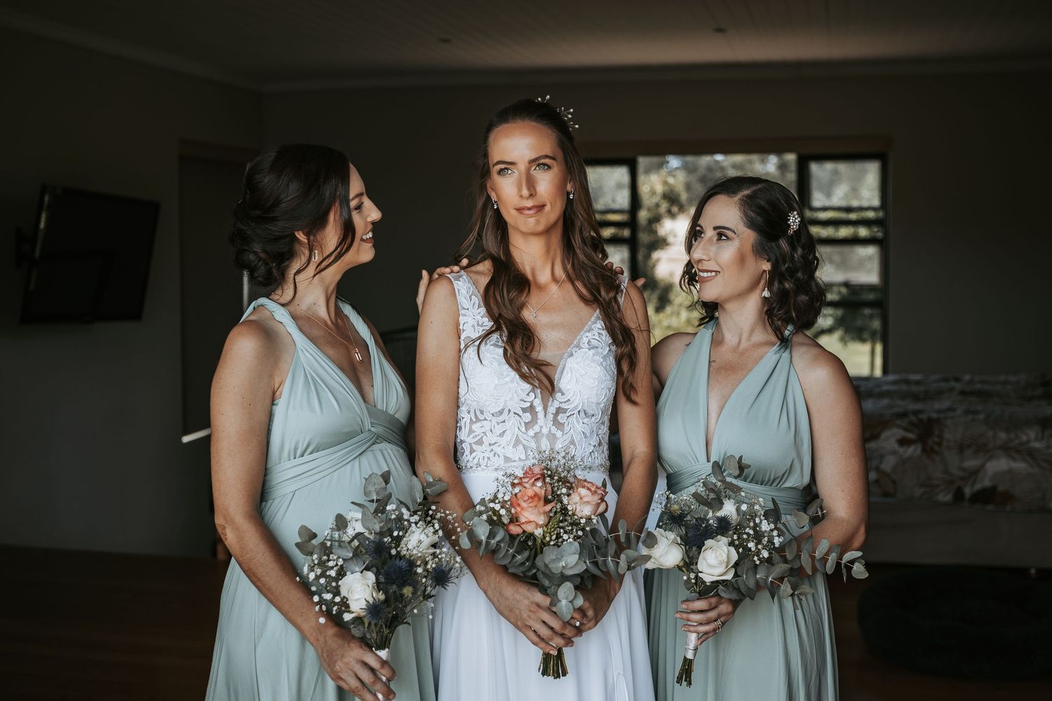 Bridesmaids in soft blue dresses hold bouquets during indoor wedding preparations.