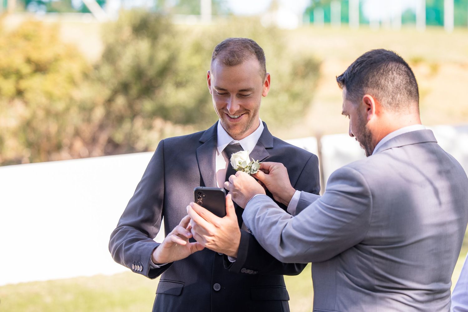 Two men in suits share a lighthearted moment outdoors during a wedding celebration.