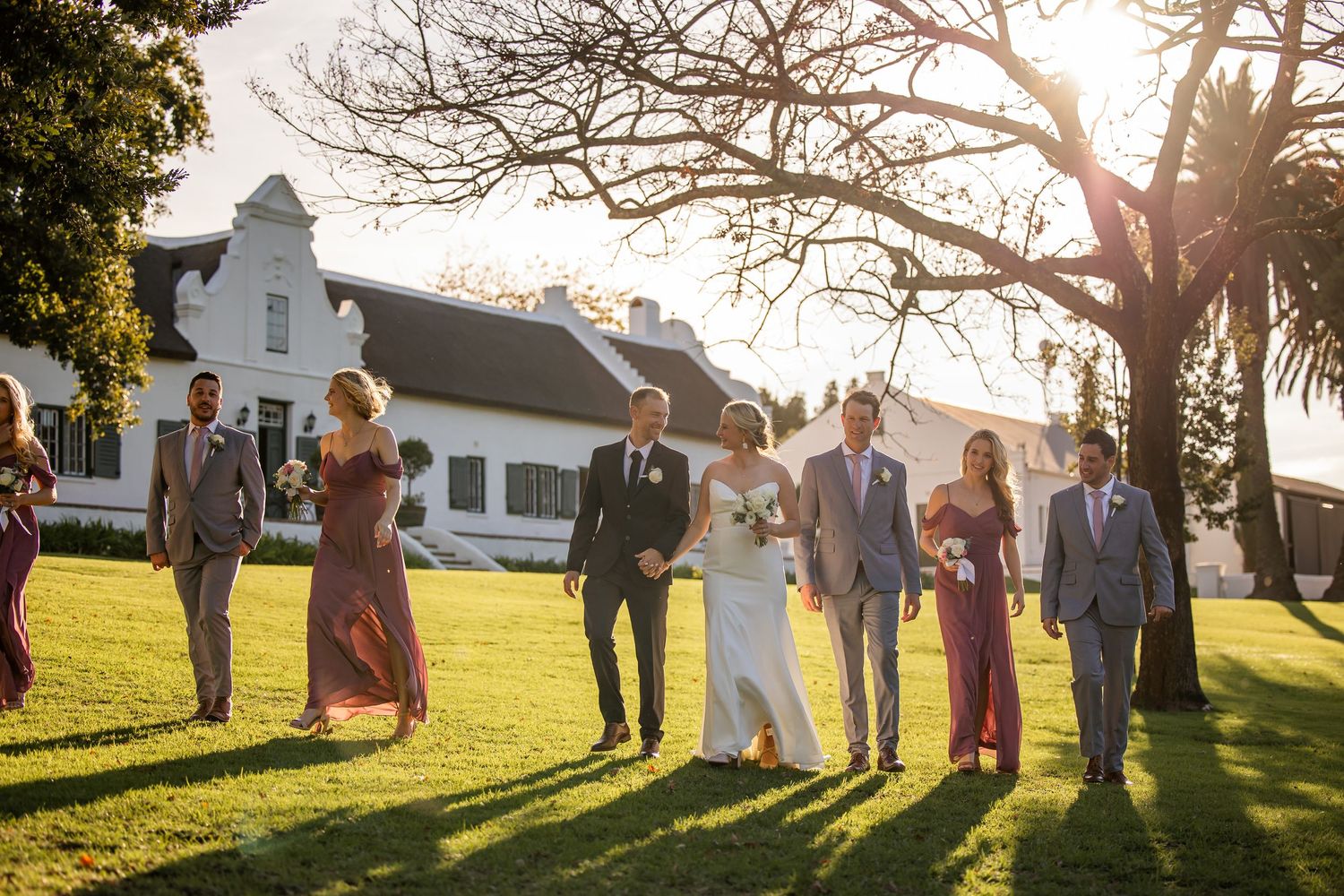 Wedding party walking together on a grassy lawn at sunset with a white house in the background.