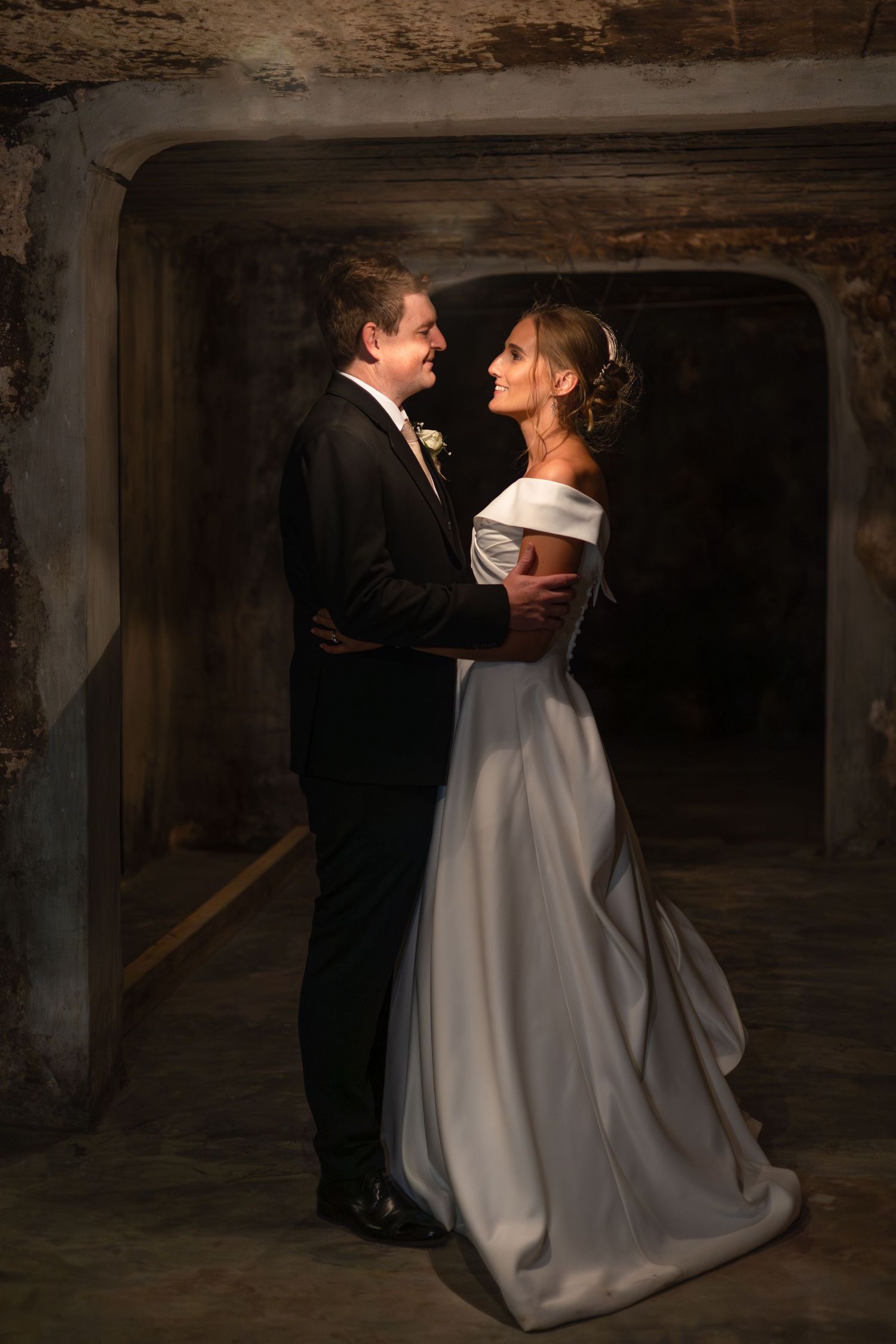Bride and groom share a romantic moment in a rustic stone cellar, the bride in an elegant off-shoulder white gown.