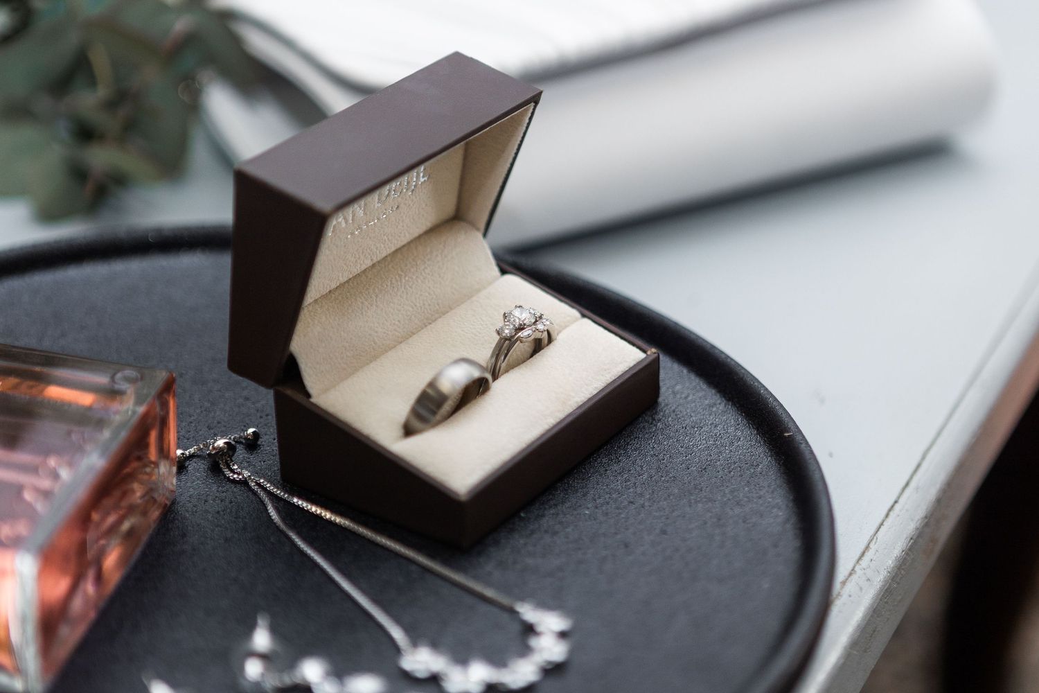 Wedding rings in a dark brown box displayed on a tray with perfume and necklace.