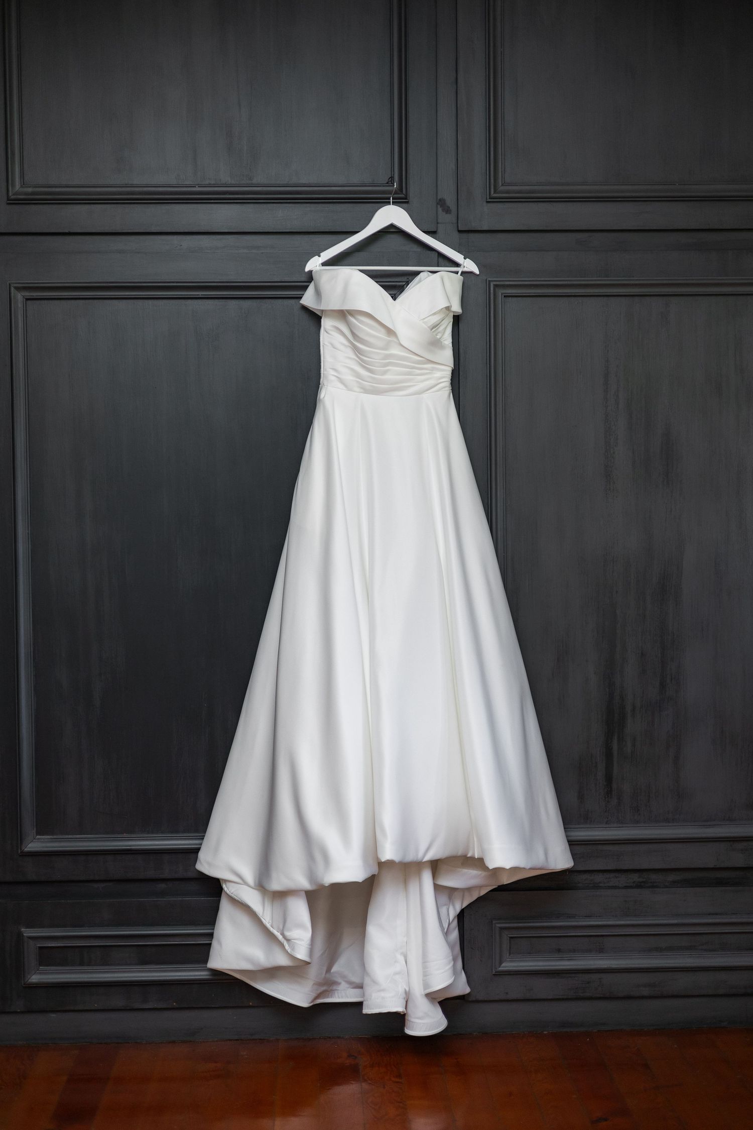Elegant off-shoulder white satin wedding dress hanging on a hanger against a dark grey paneled wall.