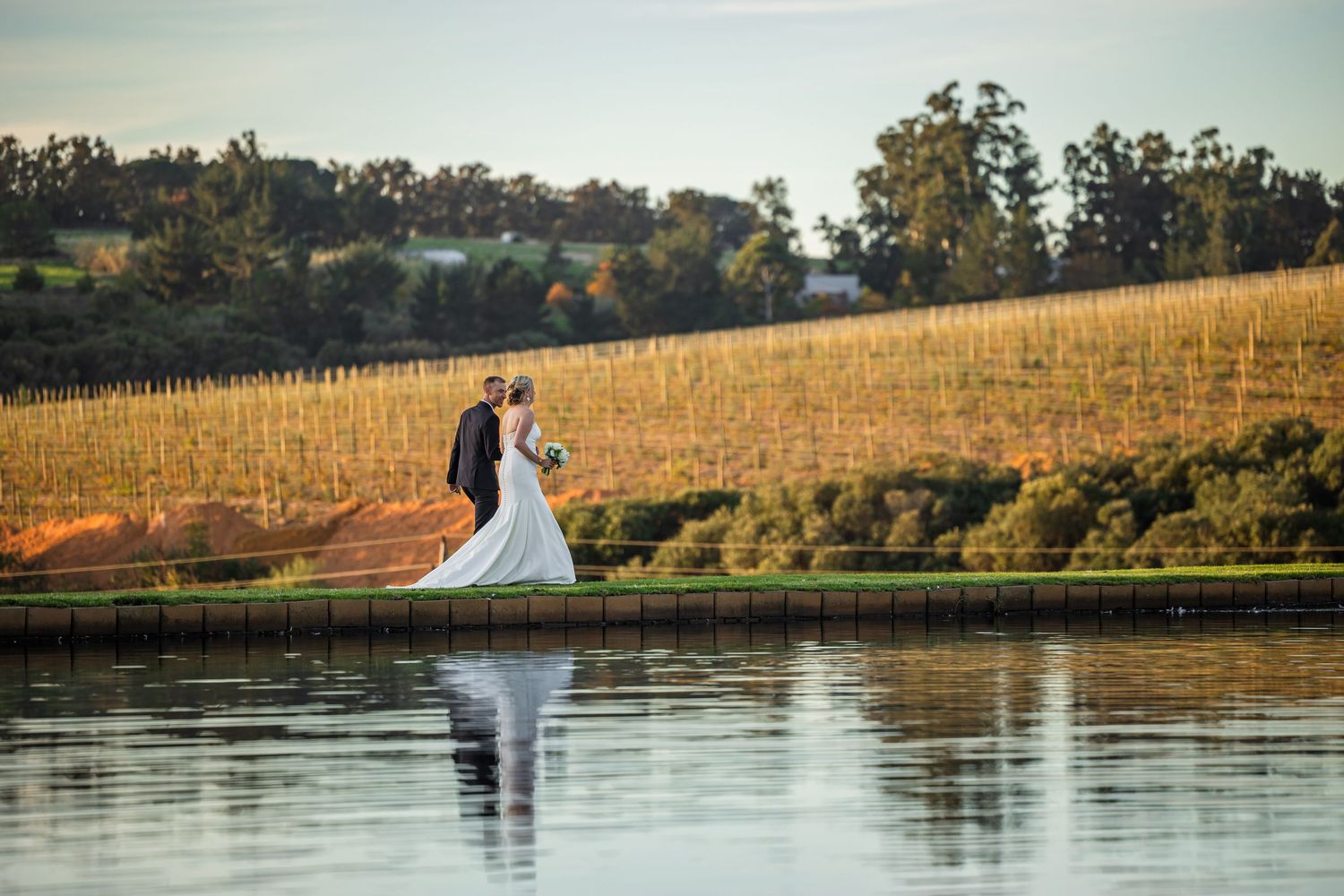 A romantic wedding couple stands by a lake at sunset with rolling vineyard hills in the background.