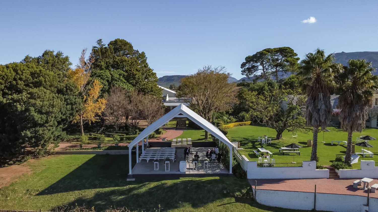 Aerial view of a scenic wedding venue with white marquee surrounded by lush gardens and mountains.