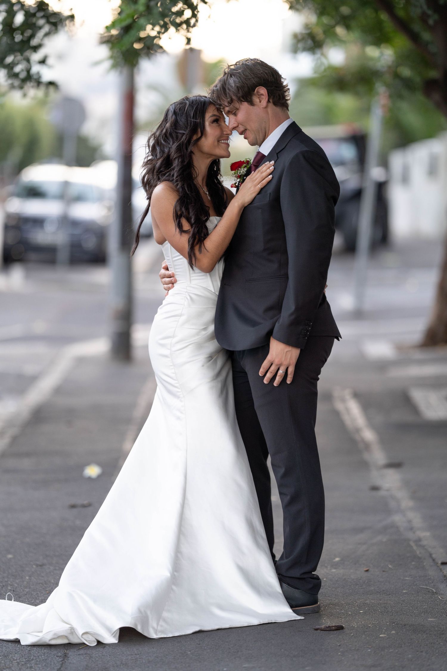 Bride and groom share a romantic embrace on a city street, wearing white gown and dark suit on their wedding day.