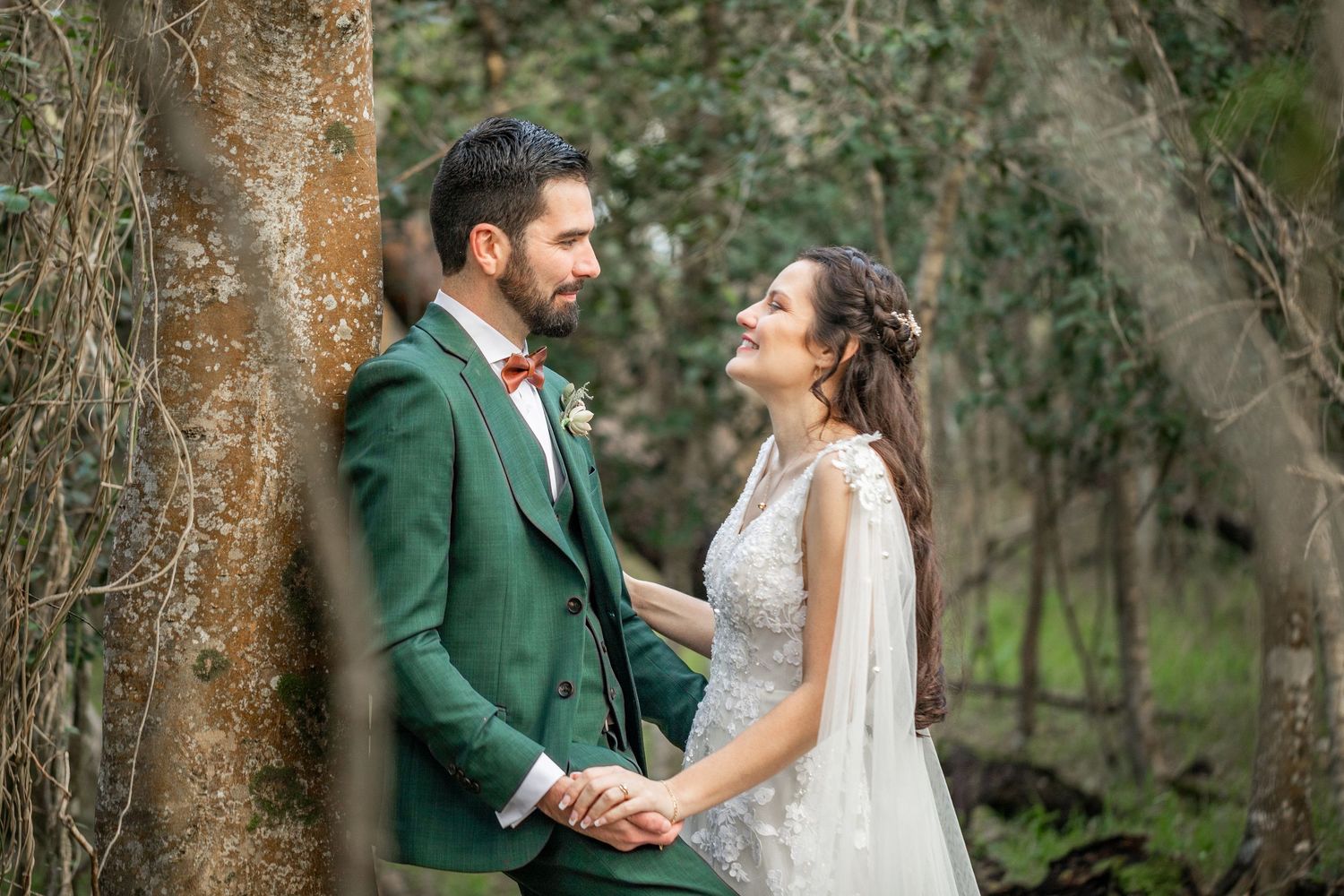 Bride and groom in green suit hold hands smiling at each other outdoors among trees in a woodland wedding setting.