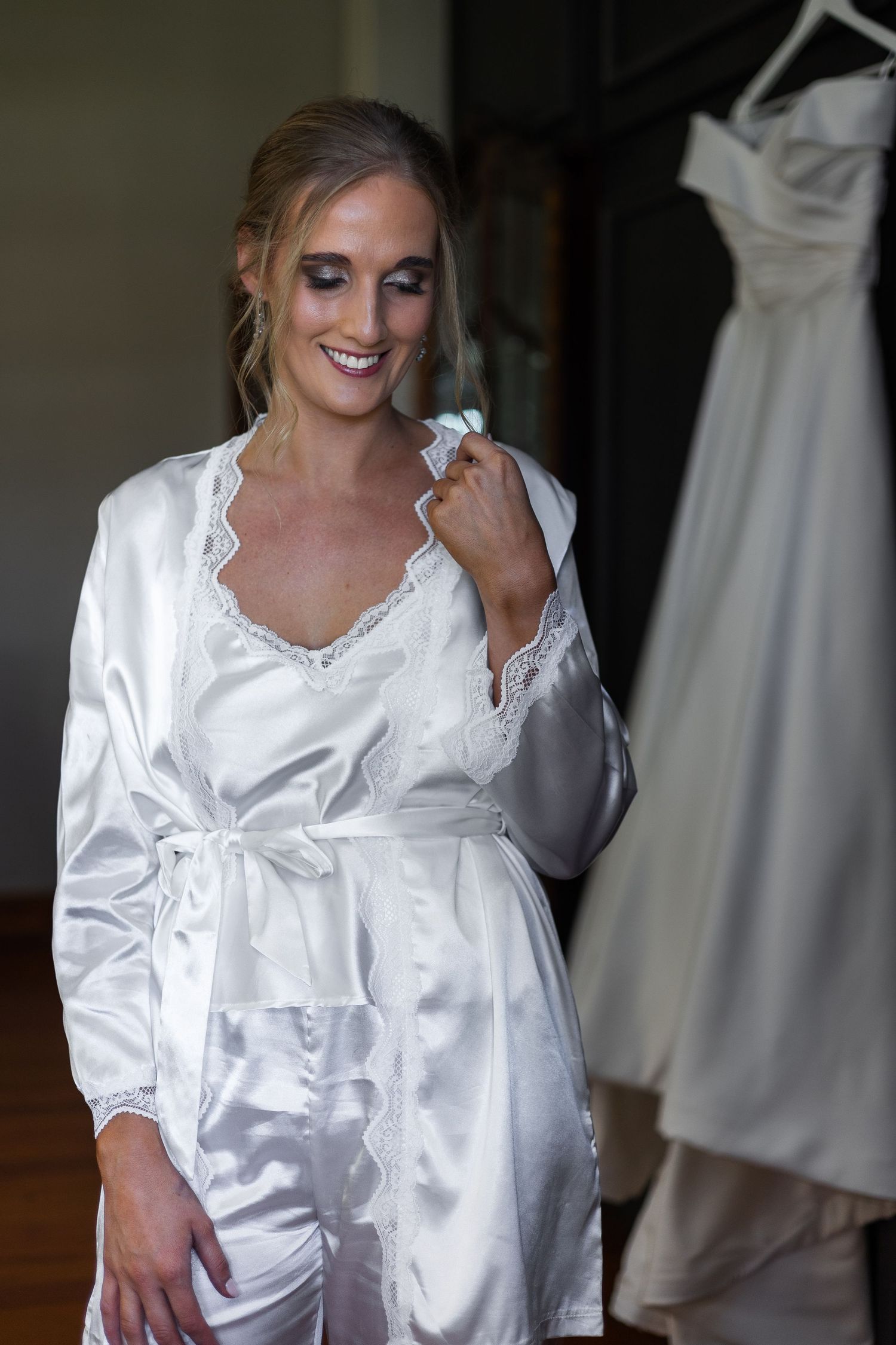 Bride in white satin lace robe smiling beside her wedding dress on a hanger.